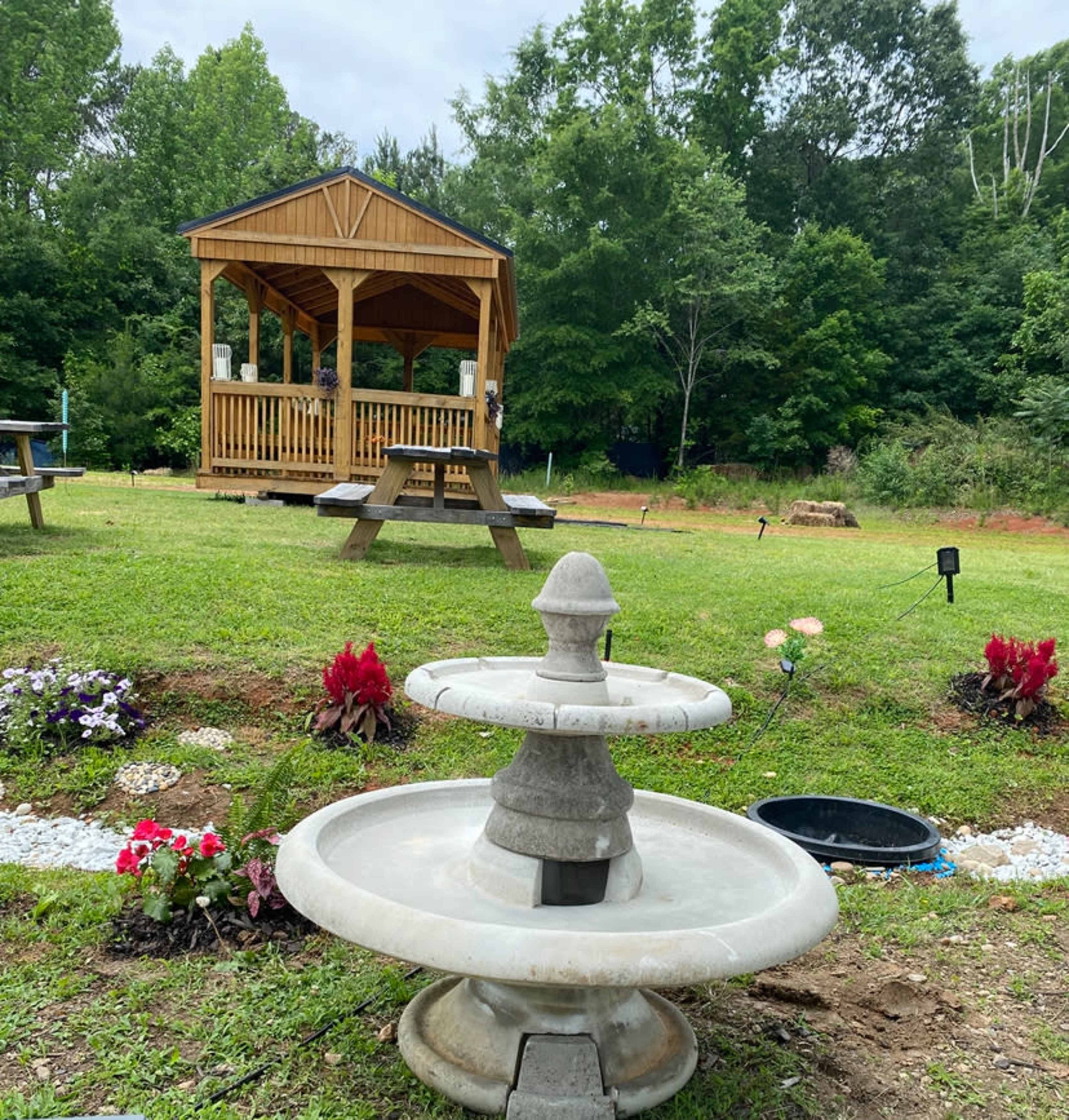 A stone fountain is situated in the foreground, while a wooden gazebo stands in the background amid a landscaped garden with various plants.