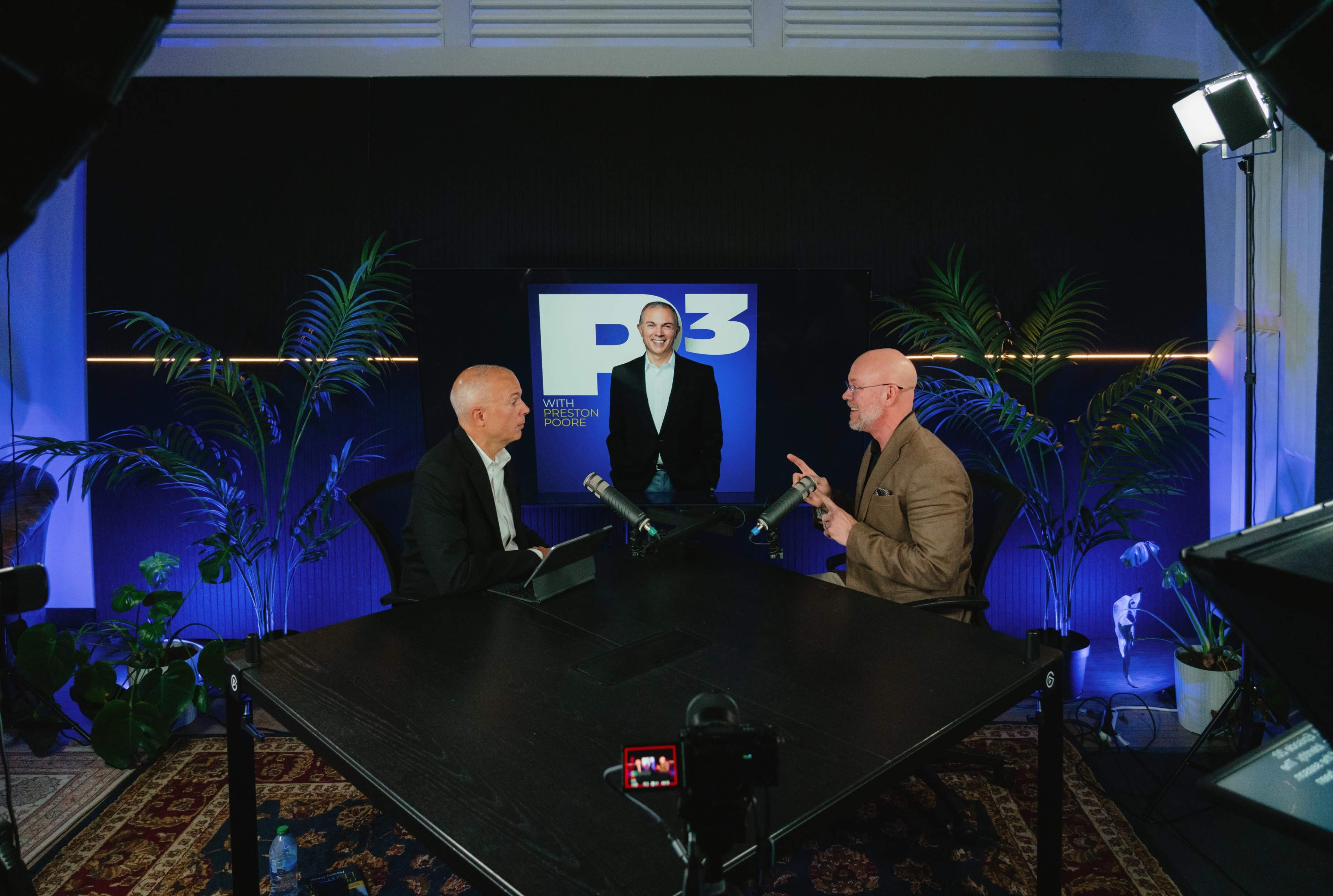 Three men are sitting at a table in a studio setting, with one man in the background standing and two men in front engaged in a discussion, while a logo on the wall displays "P3."