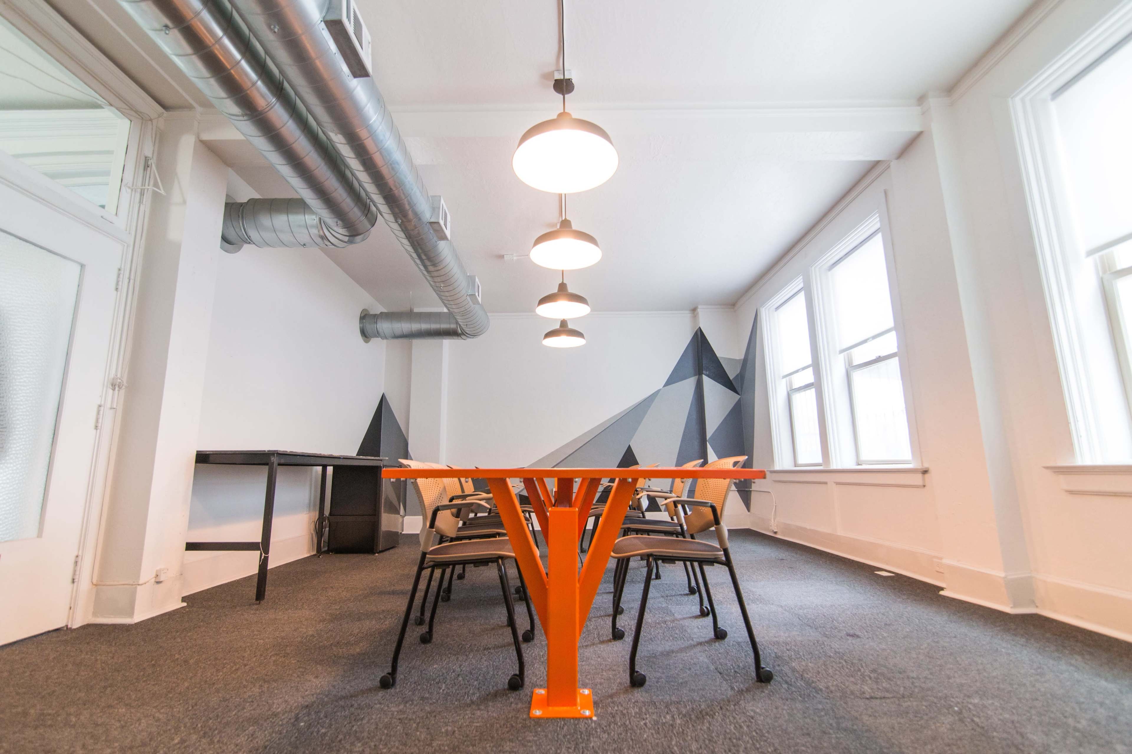 A conference room features an orange table with several chairs, complemented by overhead lights and geometric wall art.