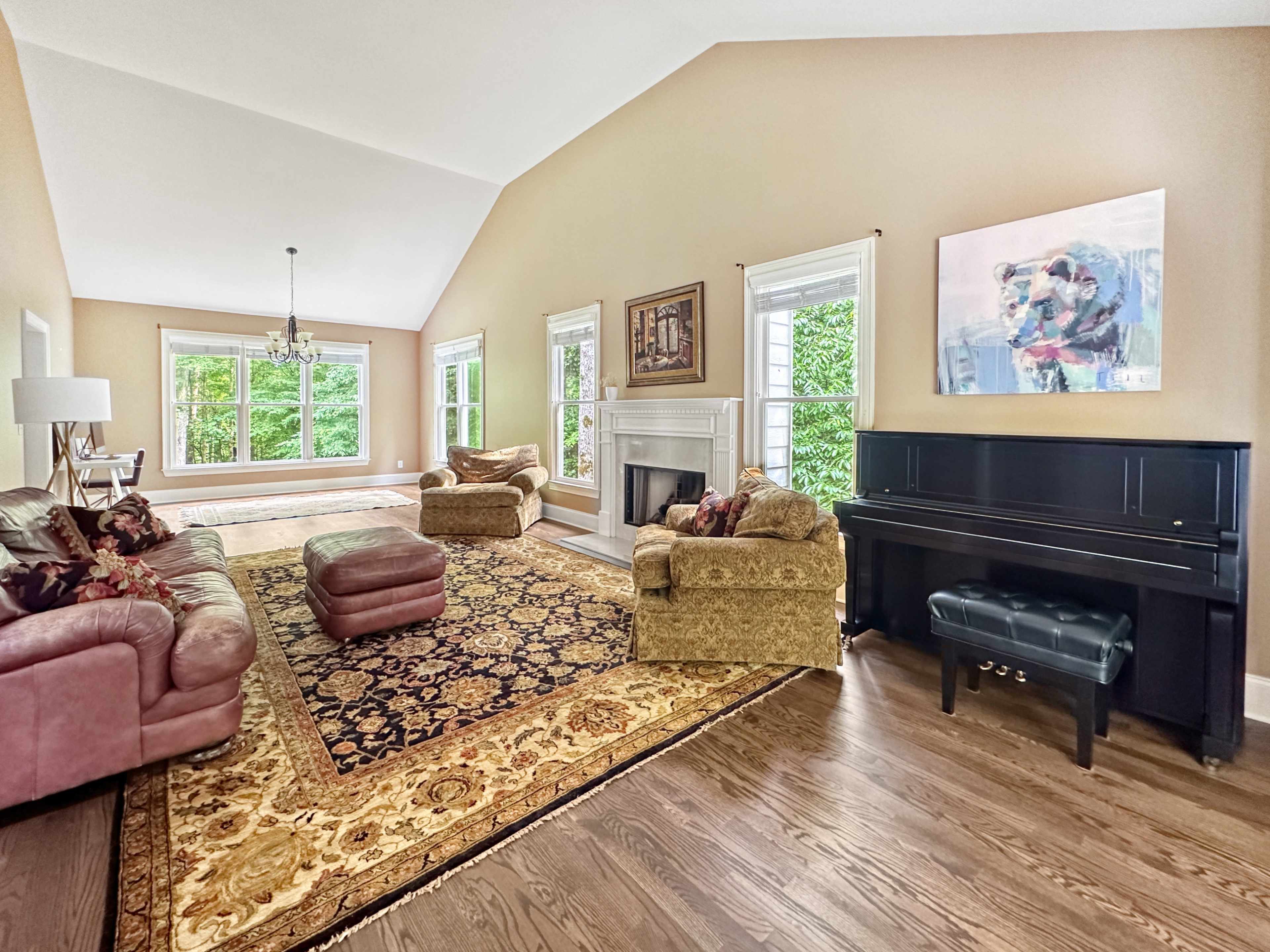 The living room features a large area rug, two sofas, a piano, and large windows that provide natural light and views of the greenery outside.