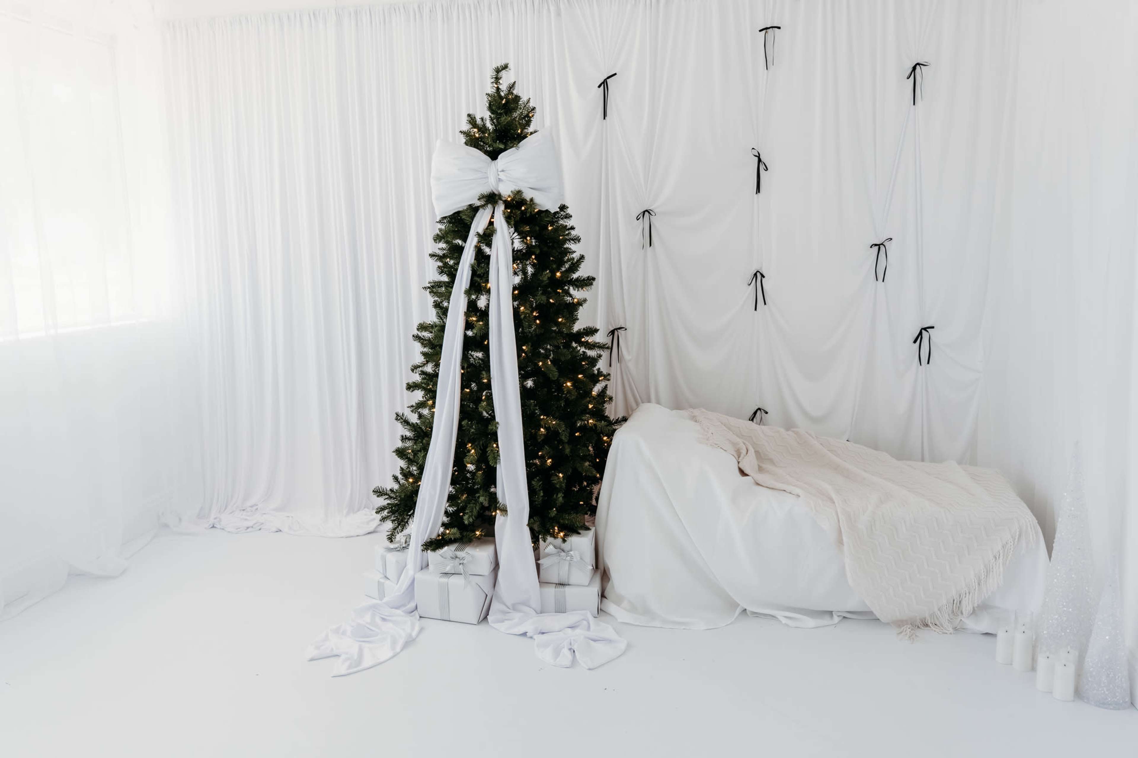 A Christmas tree with white decorations and ribbons stands next to a bed covered with a light-colored blanket in a minimalist, bright room.