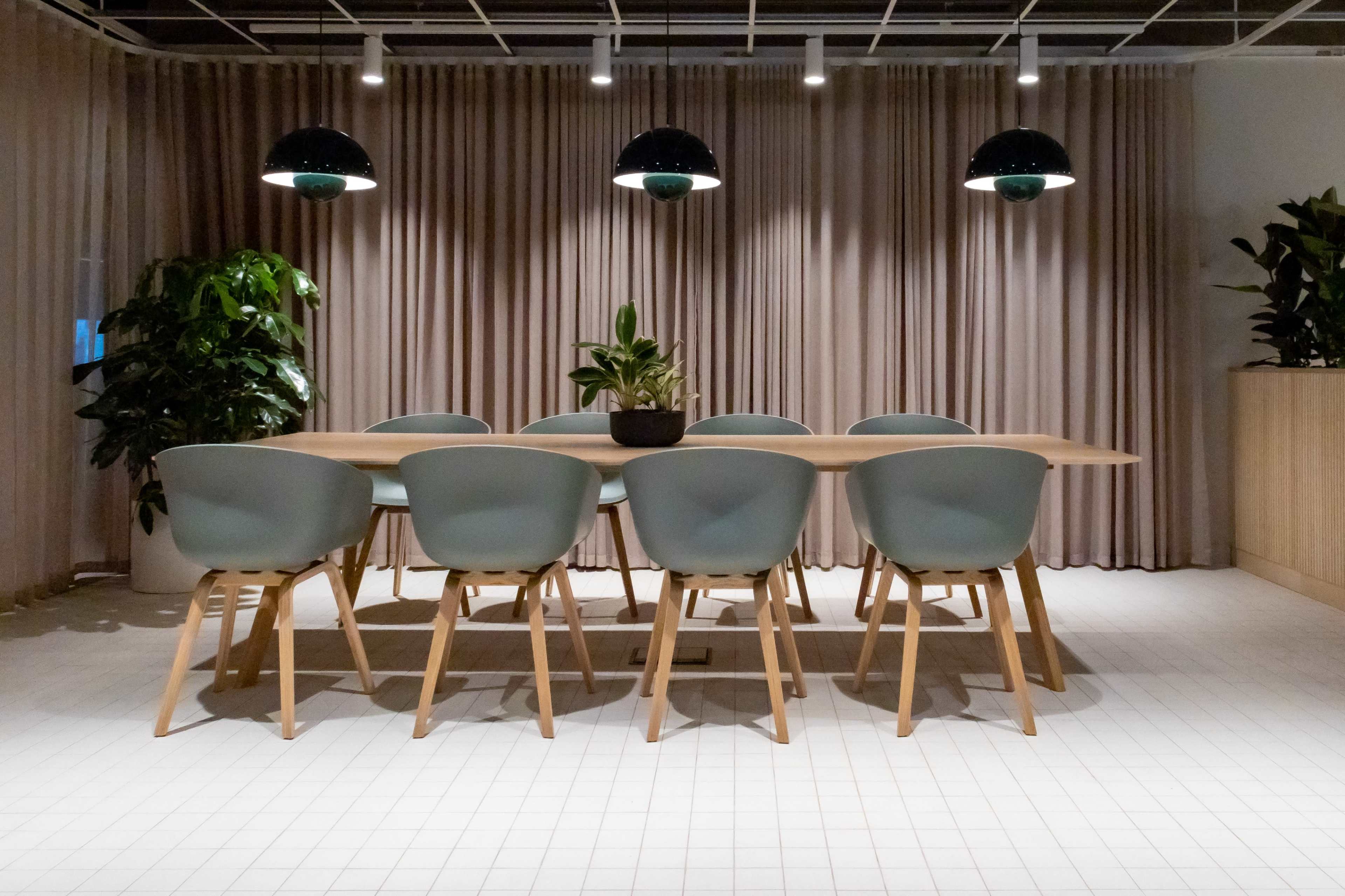 A long wooden dining table with light blue chairs is situated under black pendant lights in a room with soft beige curtains and indoor plants.