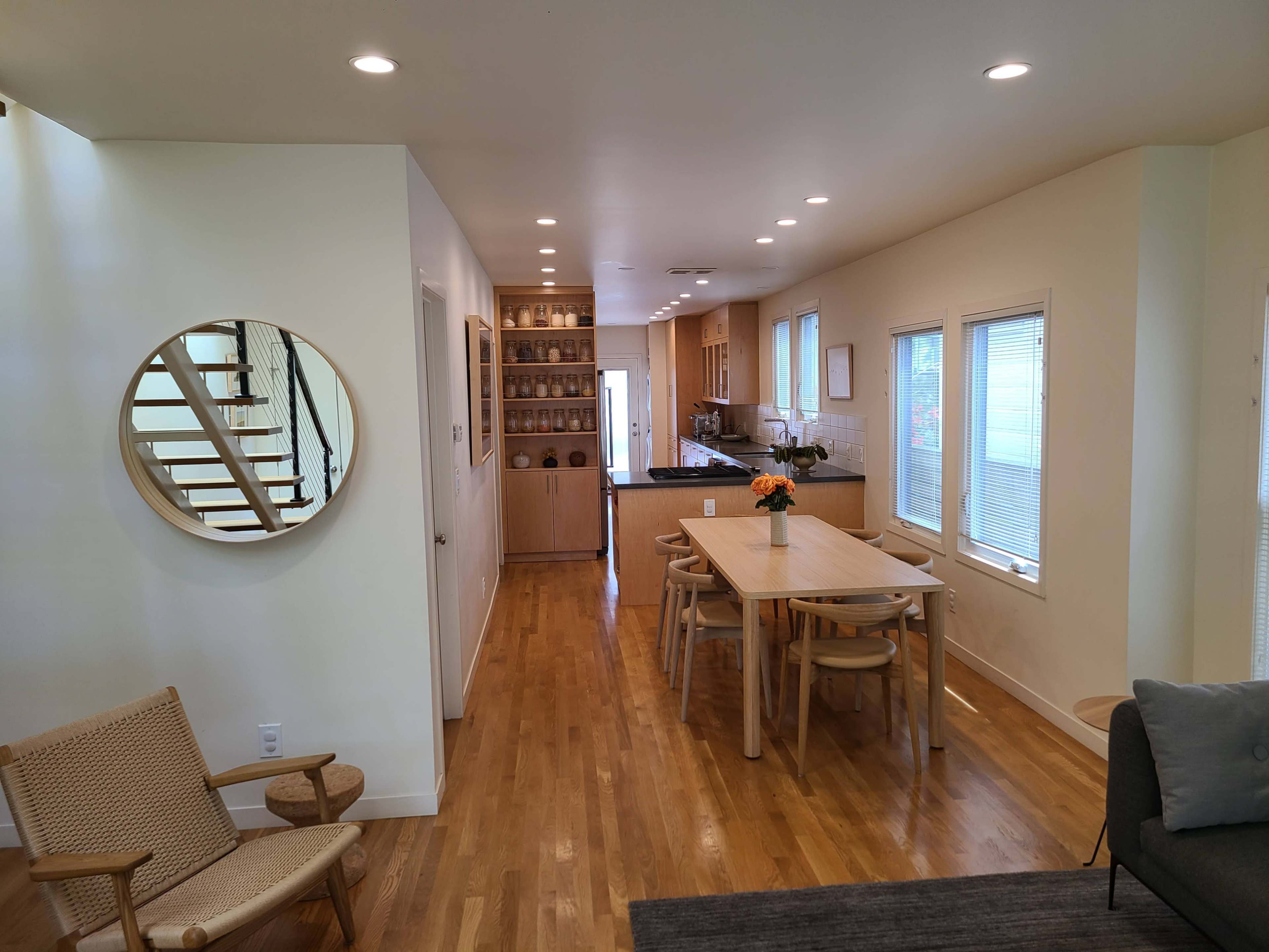 A modern interior space features a dining area with a wooden table and chairs, a kitchen in the background, and a staircase leading to the upper level.