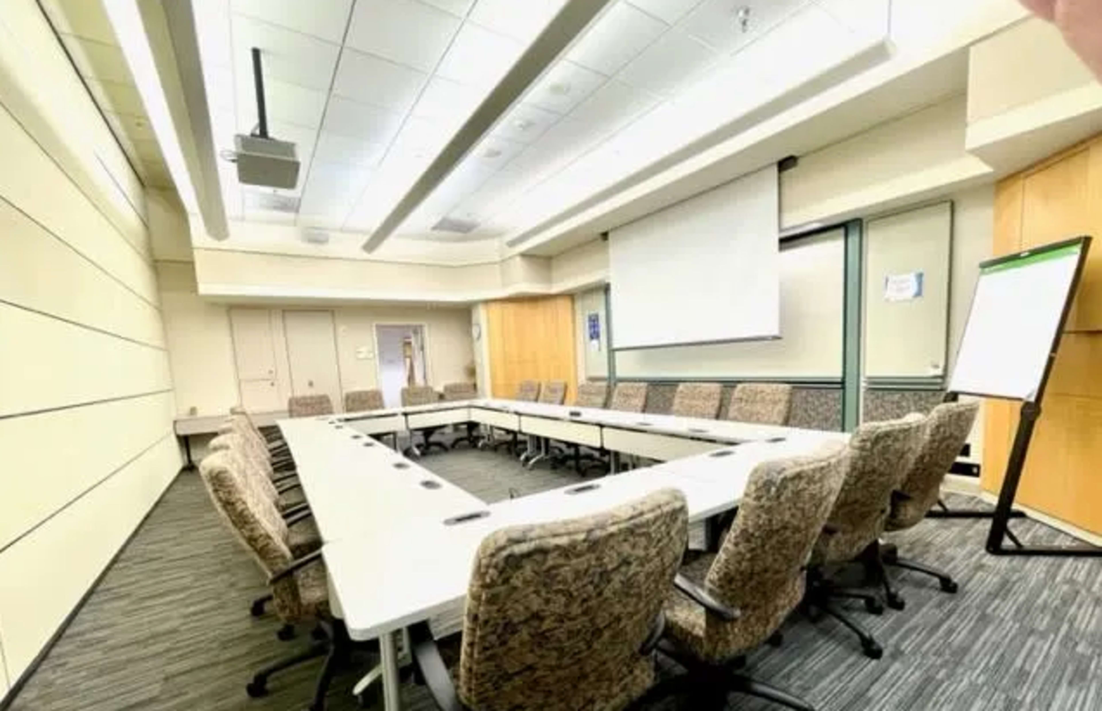 A conference room features a long table surrounded by several chairs, with a projector screen and whiteboard at one end.