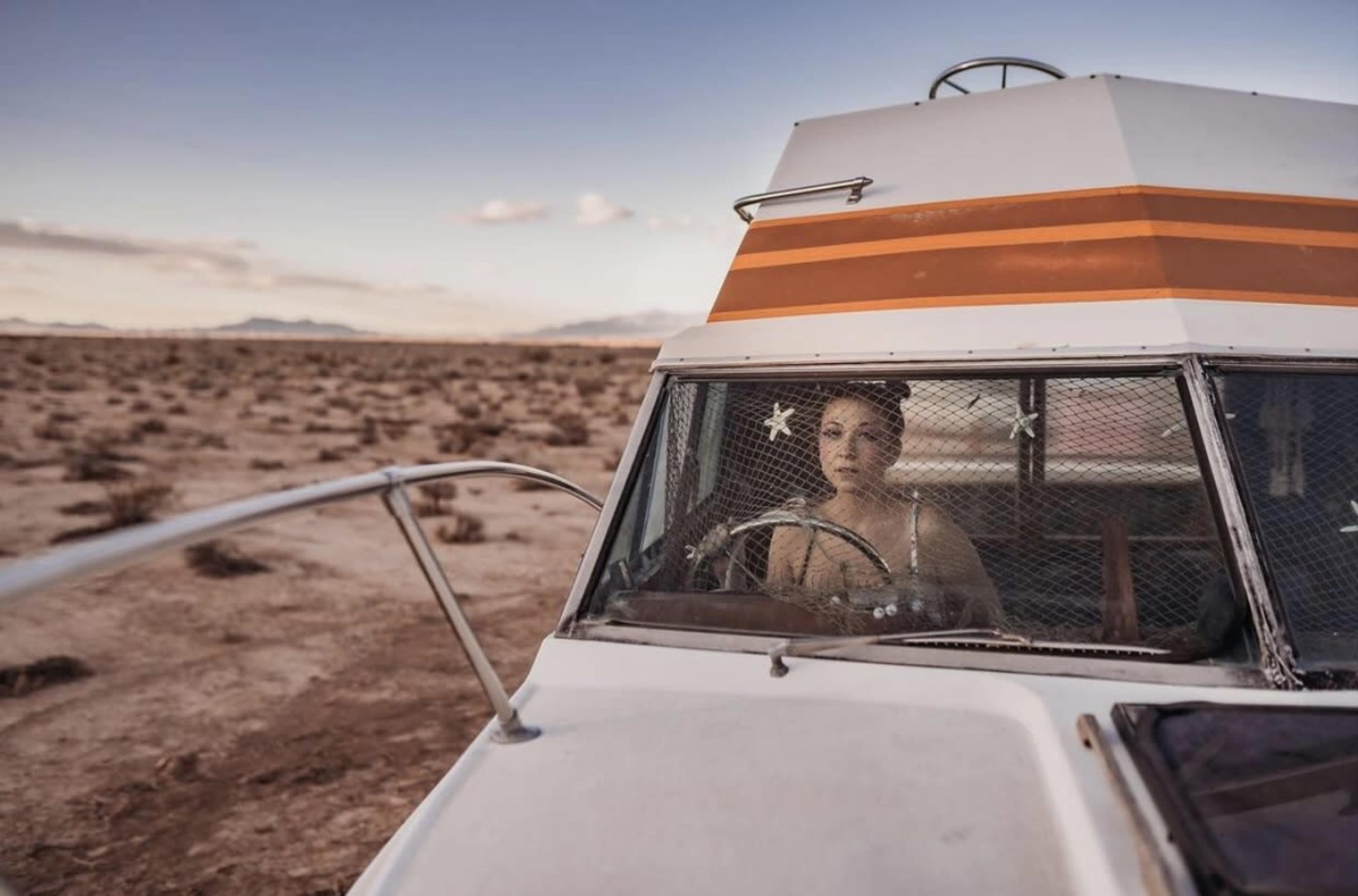 A woman sits inside a vintage camper van parked in a barren landscape with sparse vegetation and distant mountains under a clear sky.