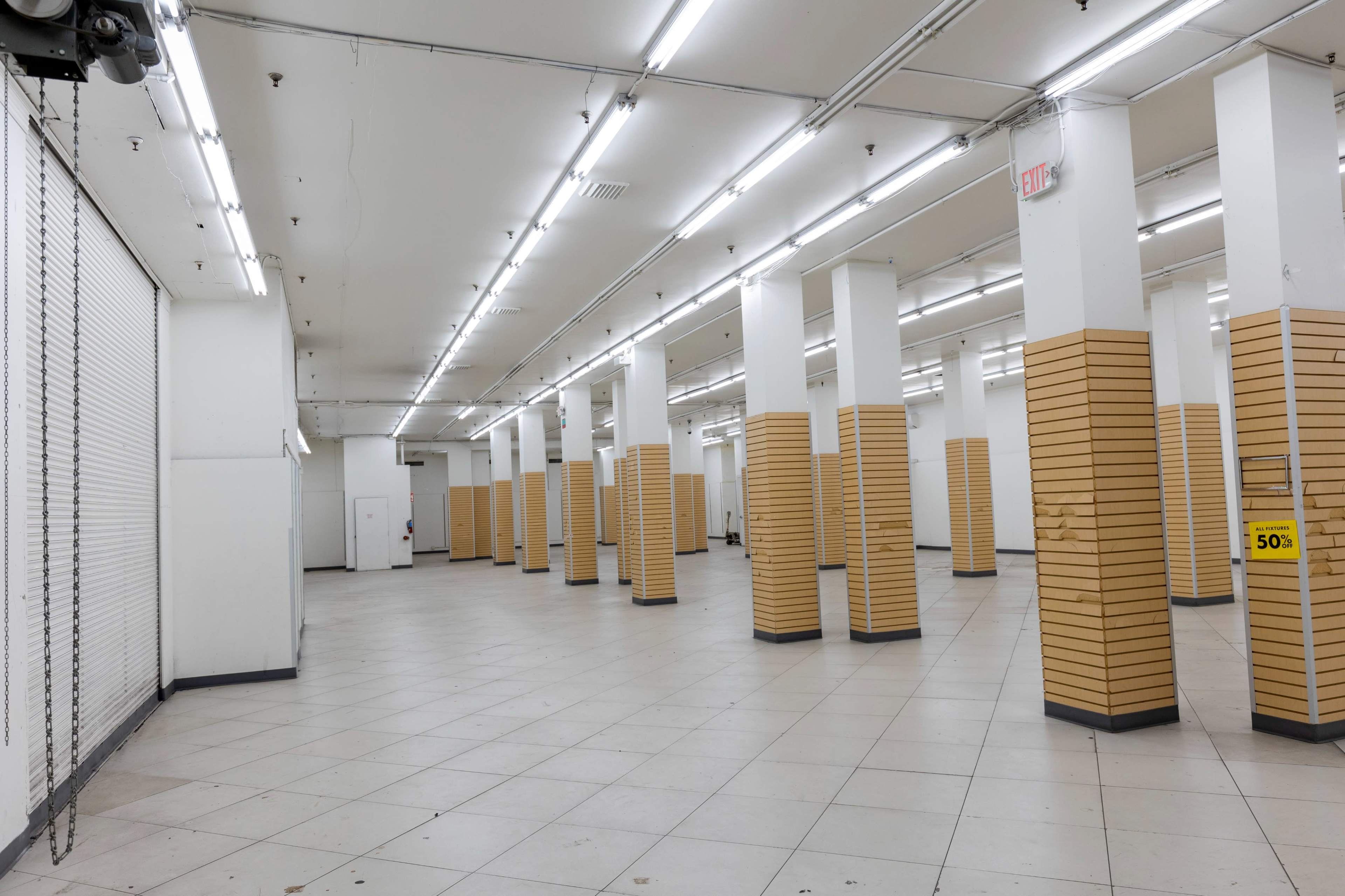 The image shows an empty retail space with white walls, fluorescent lighting, and several yellow-paneled columns.