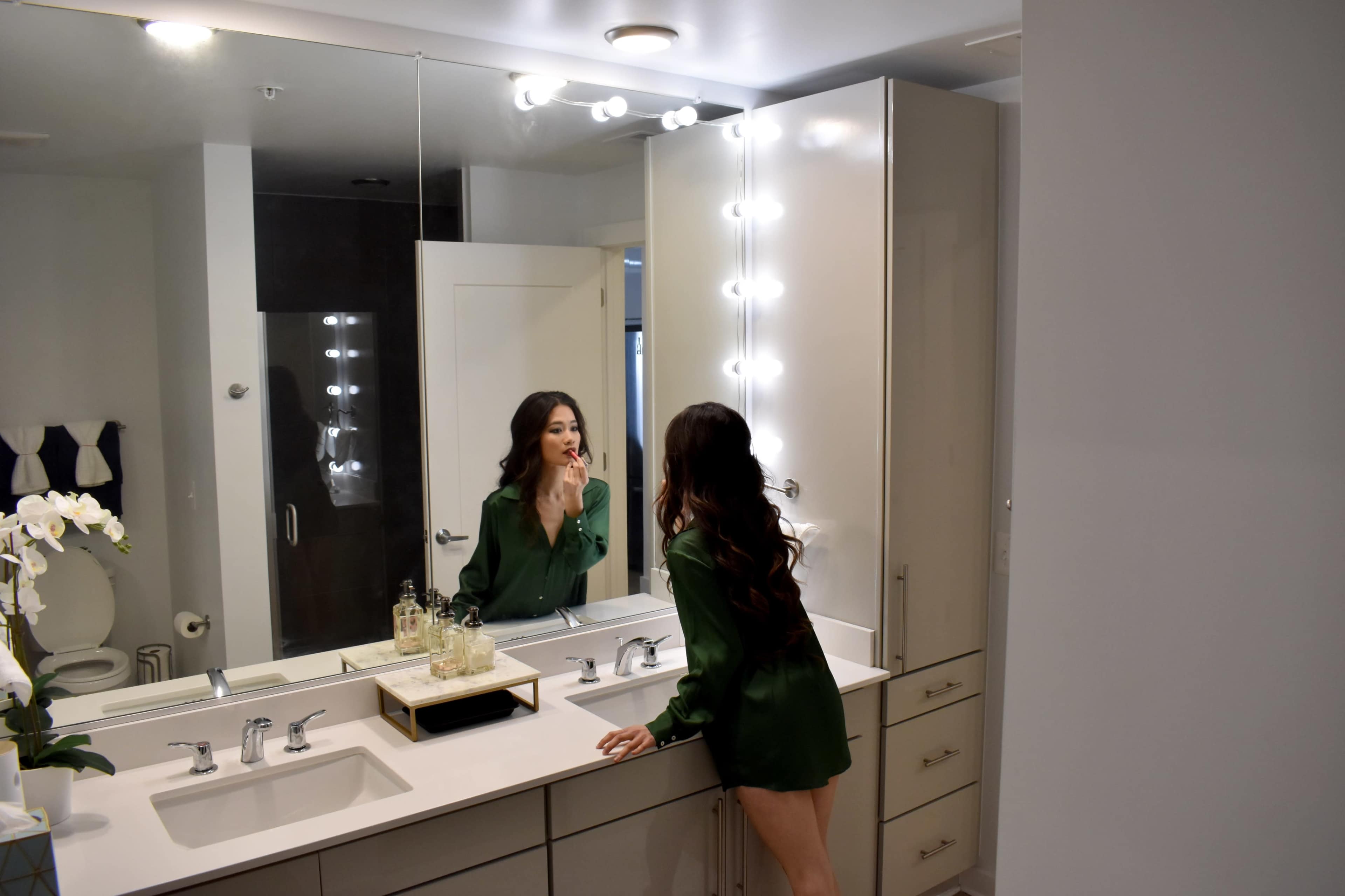 A woman in a green shirt is applying makeup in a well-lit bathroom mirror with a modern design.