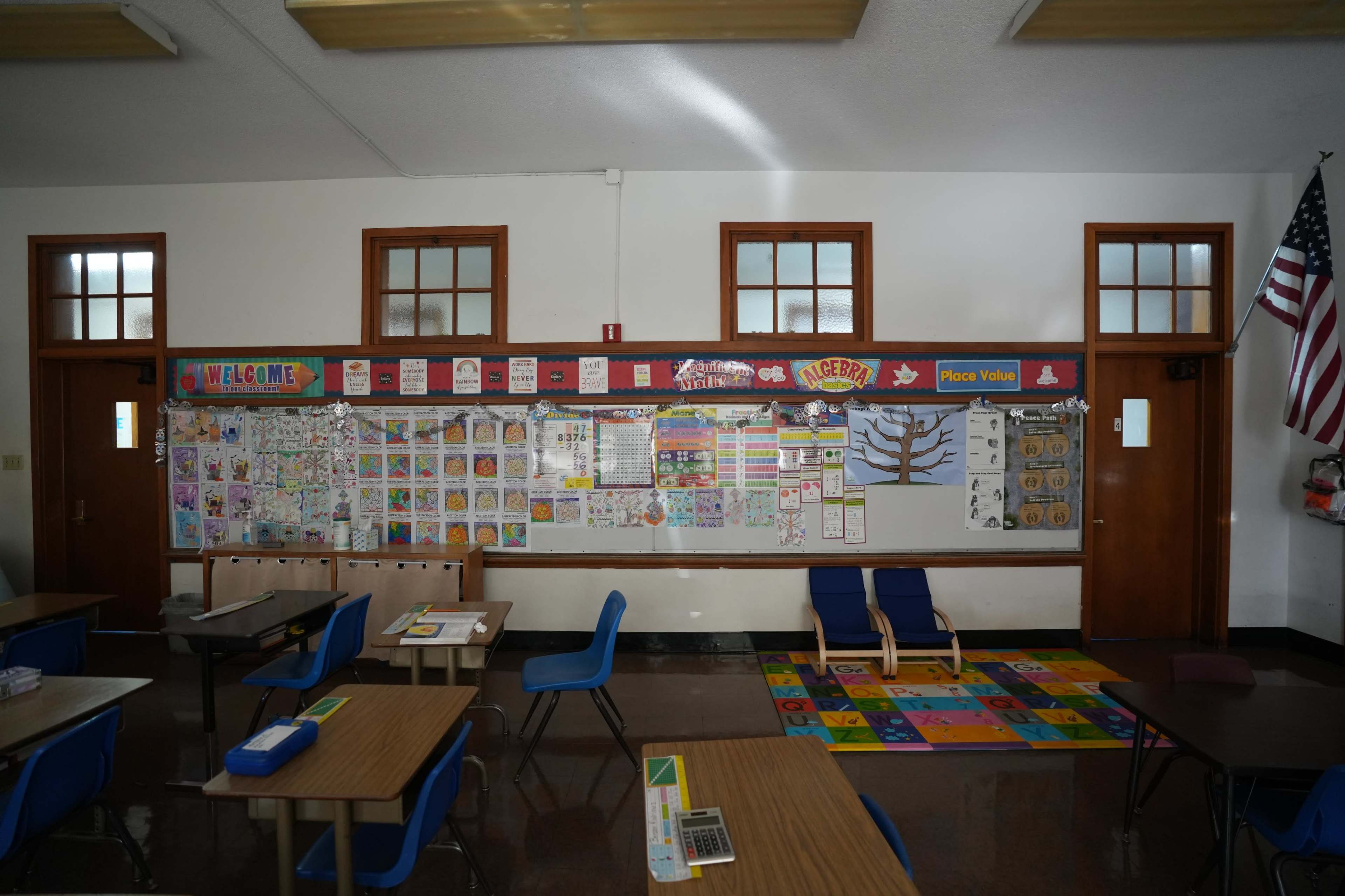 A brightly lit classroom features colorful educational posters along a bulletin board, wooden desks, and a patterned carpet.