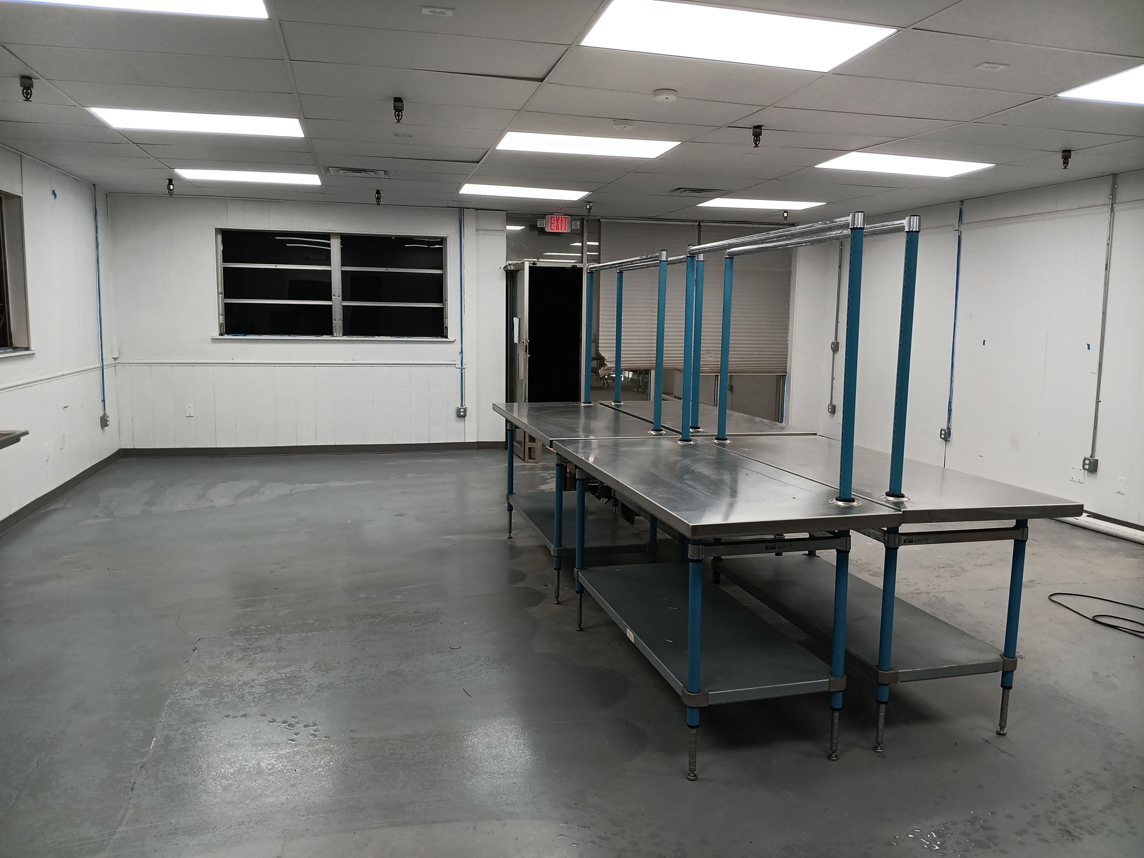 The image shows a spacious, empty room with a stainless steel worktable and shelves in the center, illuminated by overhead fluorescent lights.