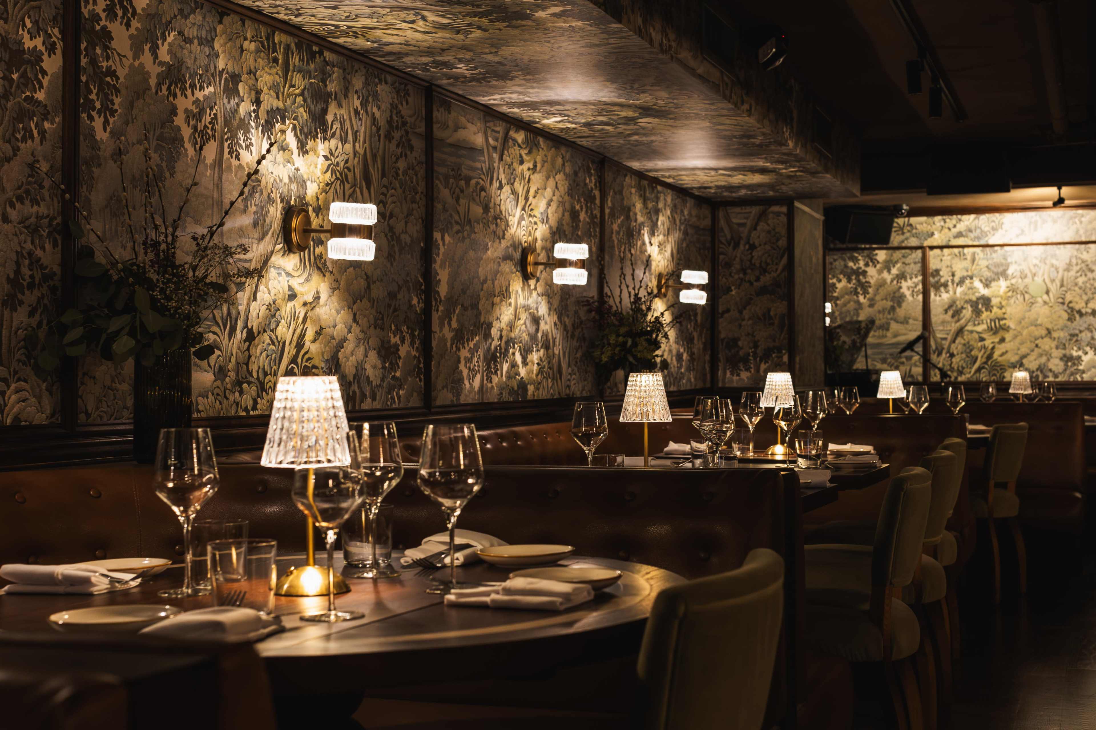 A dimly lit restaurant featuring elegant table settings with glassware and lamps, surrounded by ornate wallpaper and decorative wall sconces.