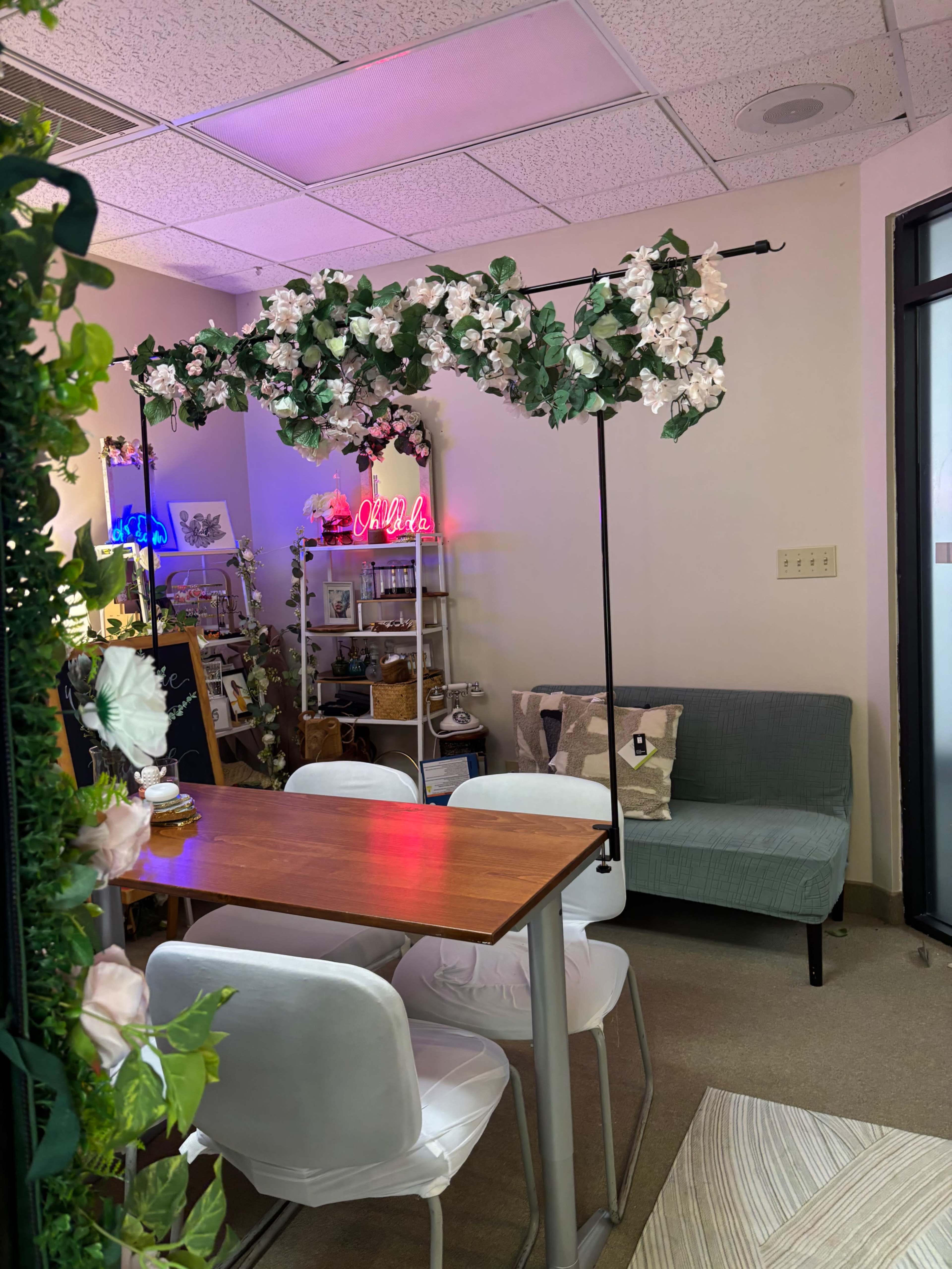 The image shows a small room featuring a wooden table with four white chairs, a green couch, and decorative plants hanging above, along with shelves displaying various items.