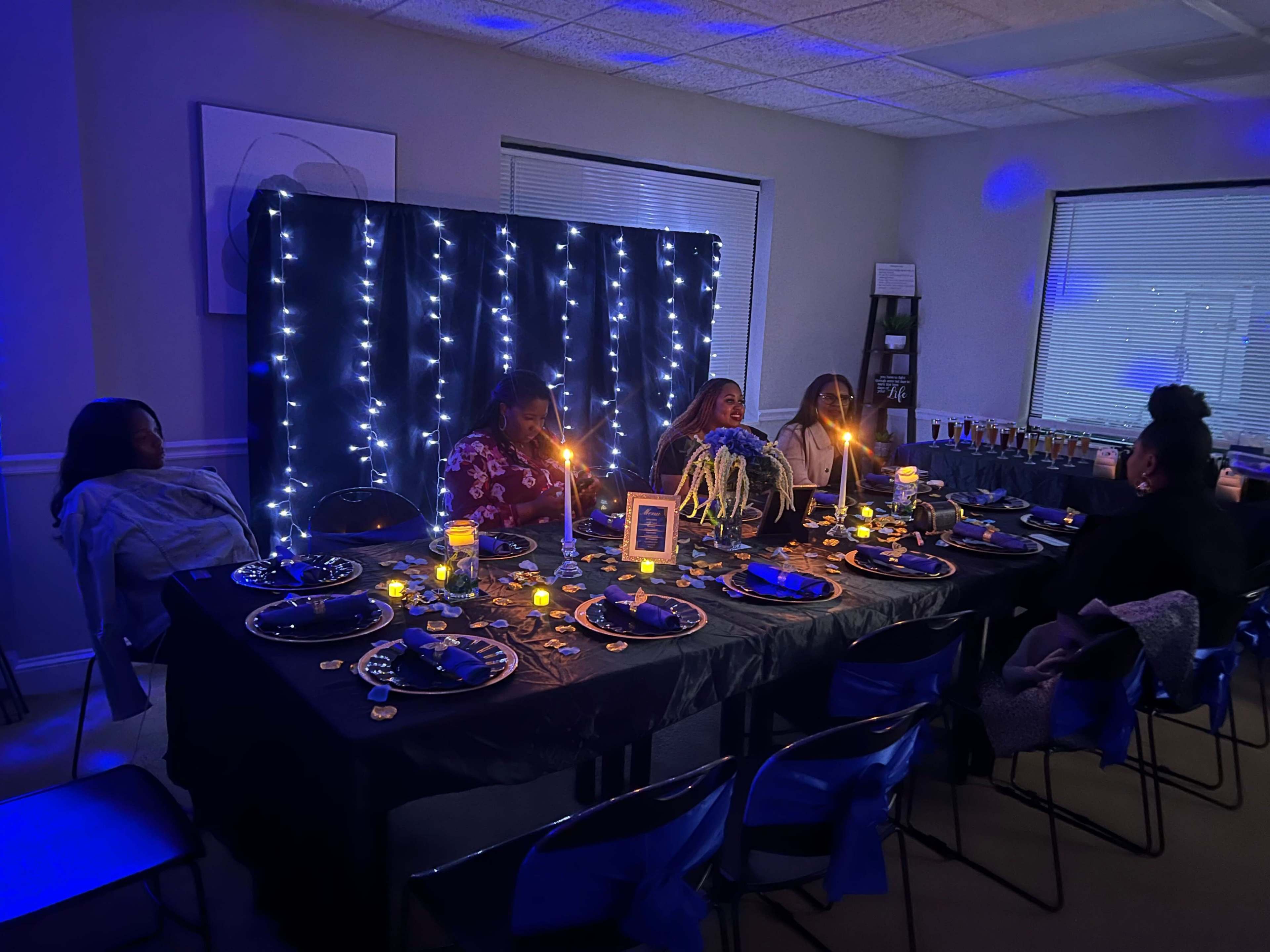 A dimly lit room features a dinner table set for five, adorned with candles and blue fairy lights in the background.