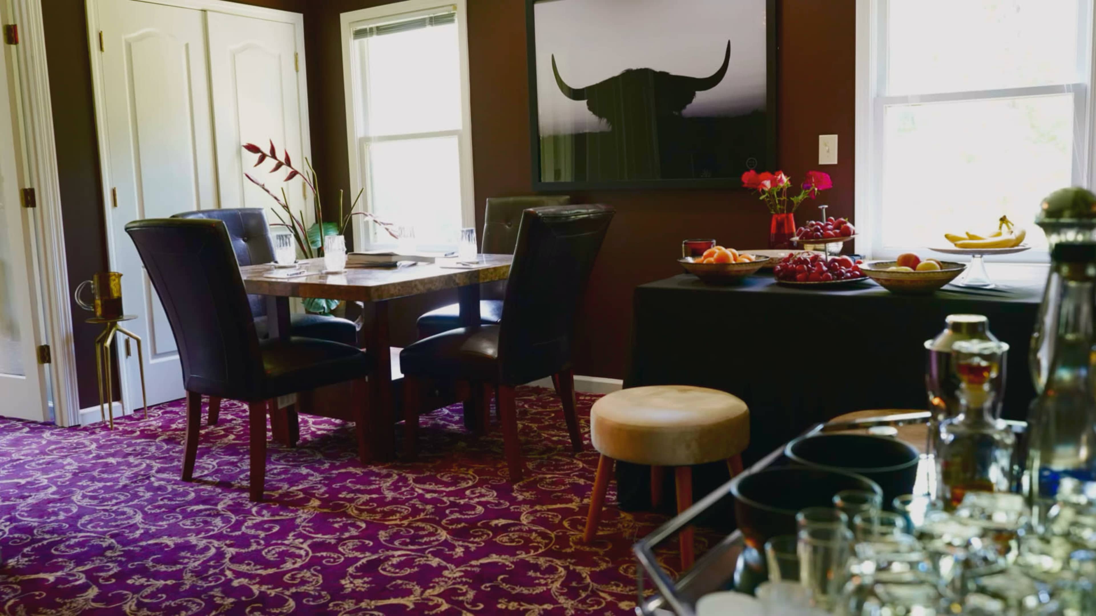 A dining area with a dark wooden table surrounded by black chairs, a floral arrangement, and a side table displaying various fruits and beverages.
