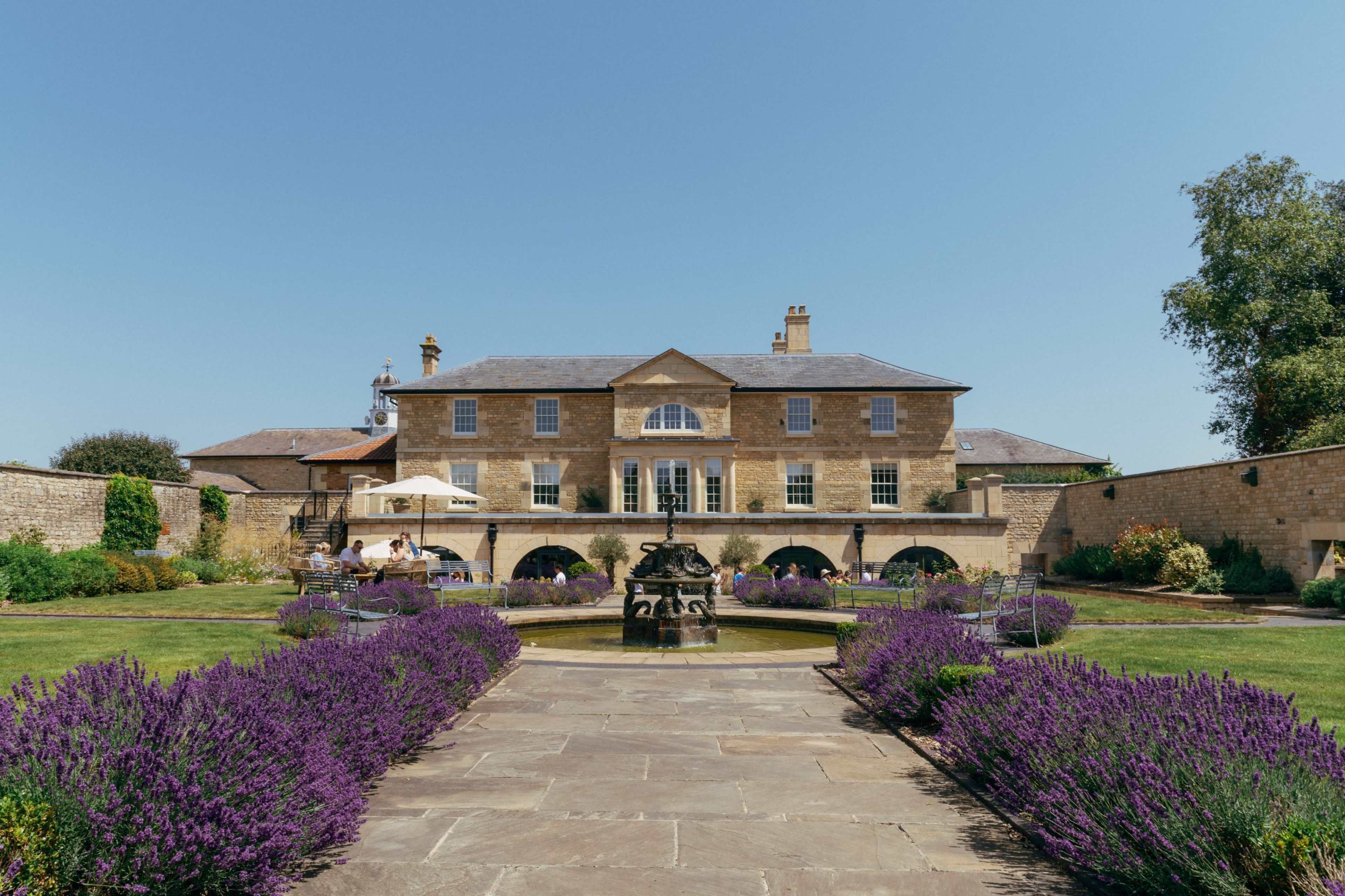 A large stone house with multiple windows and a front terrace is set against a clear blue sky, surrounded by a well-manicured garden featuring purple lavender and a central fountain.