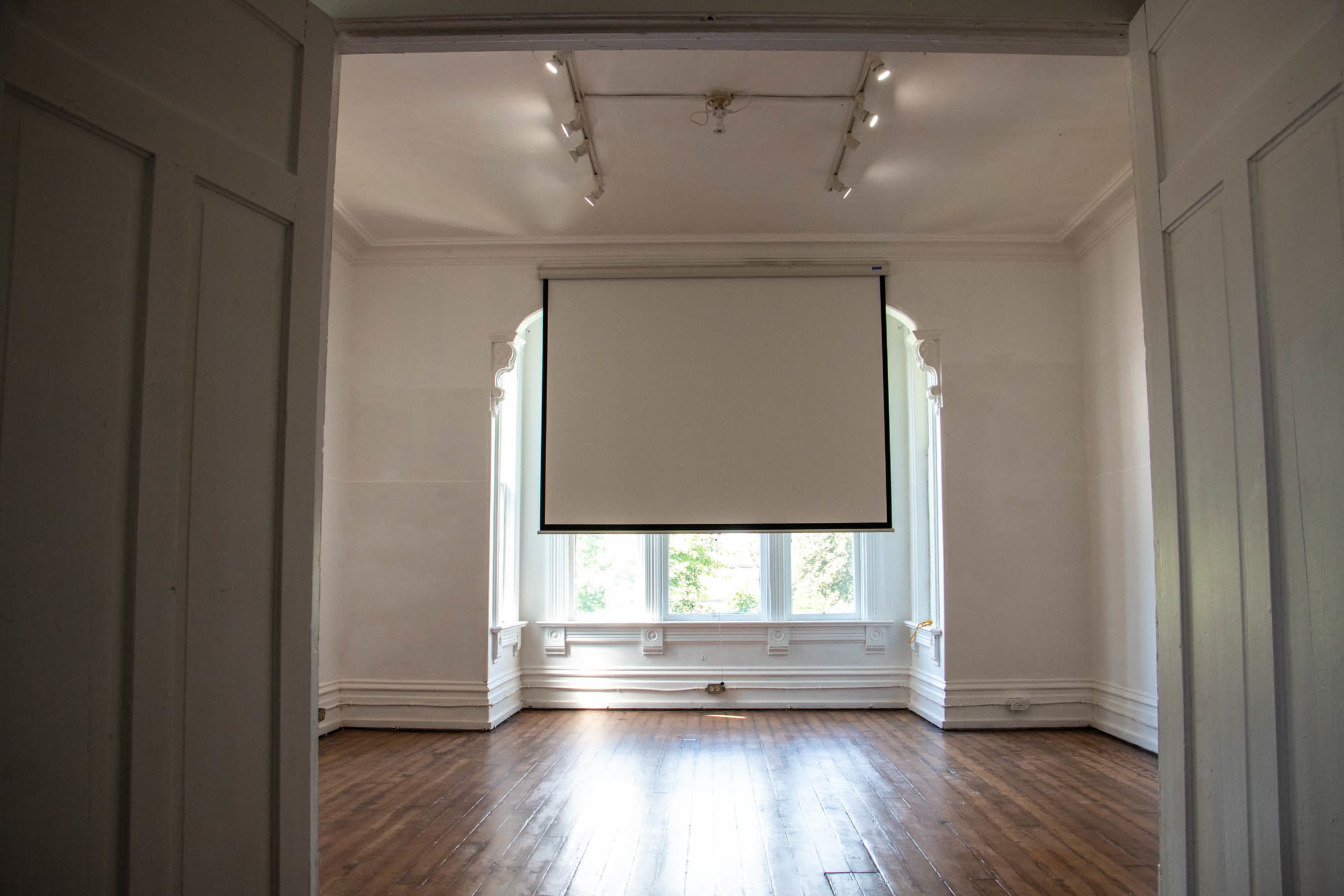 The image shows an empty room with wooden floors, a blank projection screen hanging from the ceiling, and large windows letting in natural light.