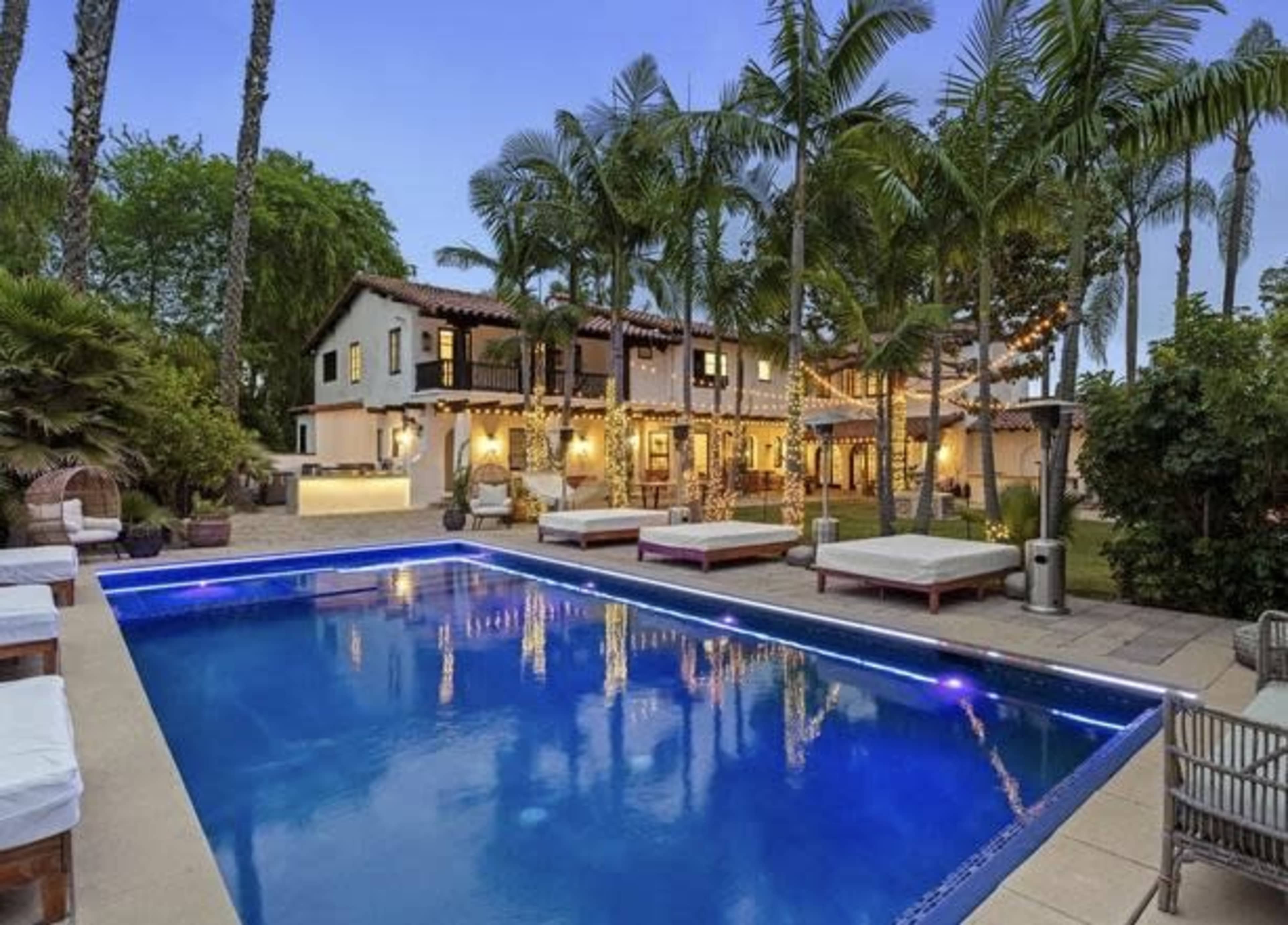 A spacious backyard features a swimming pool surrounded by lounge chairs and a two-story house adorned with palm trees and outdoor lighting.