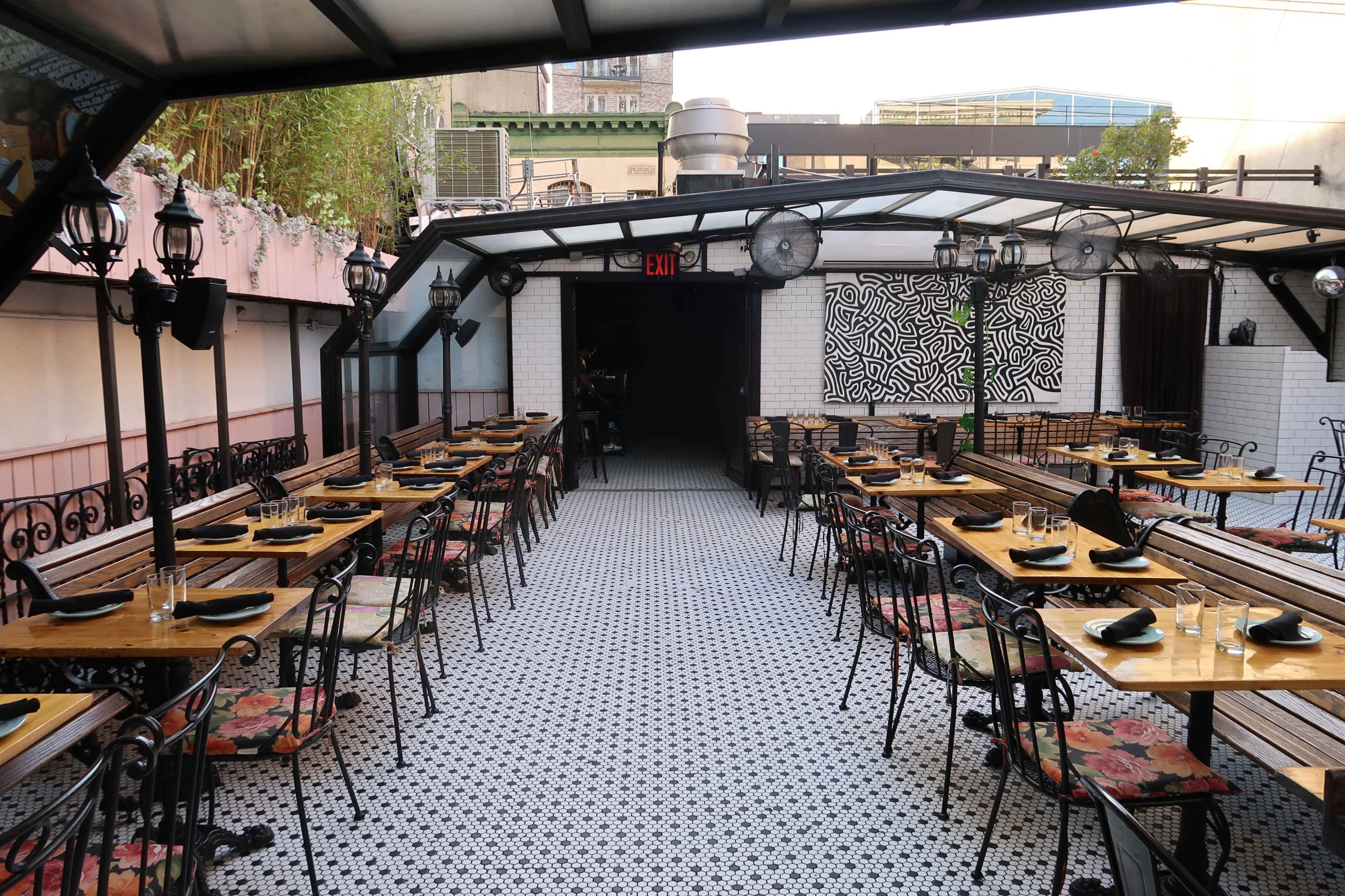 A rooftop restaurant with rows of wooden tables and chairs arranged under a metal canopy, featuring a patterned floor and decorative wall art.