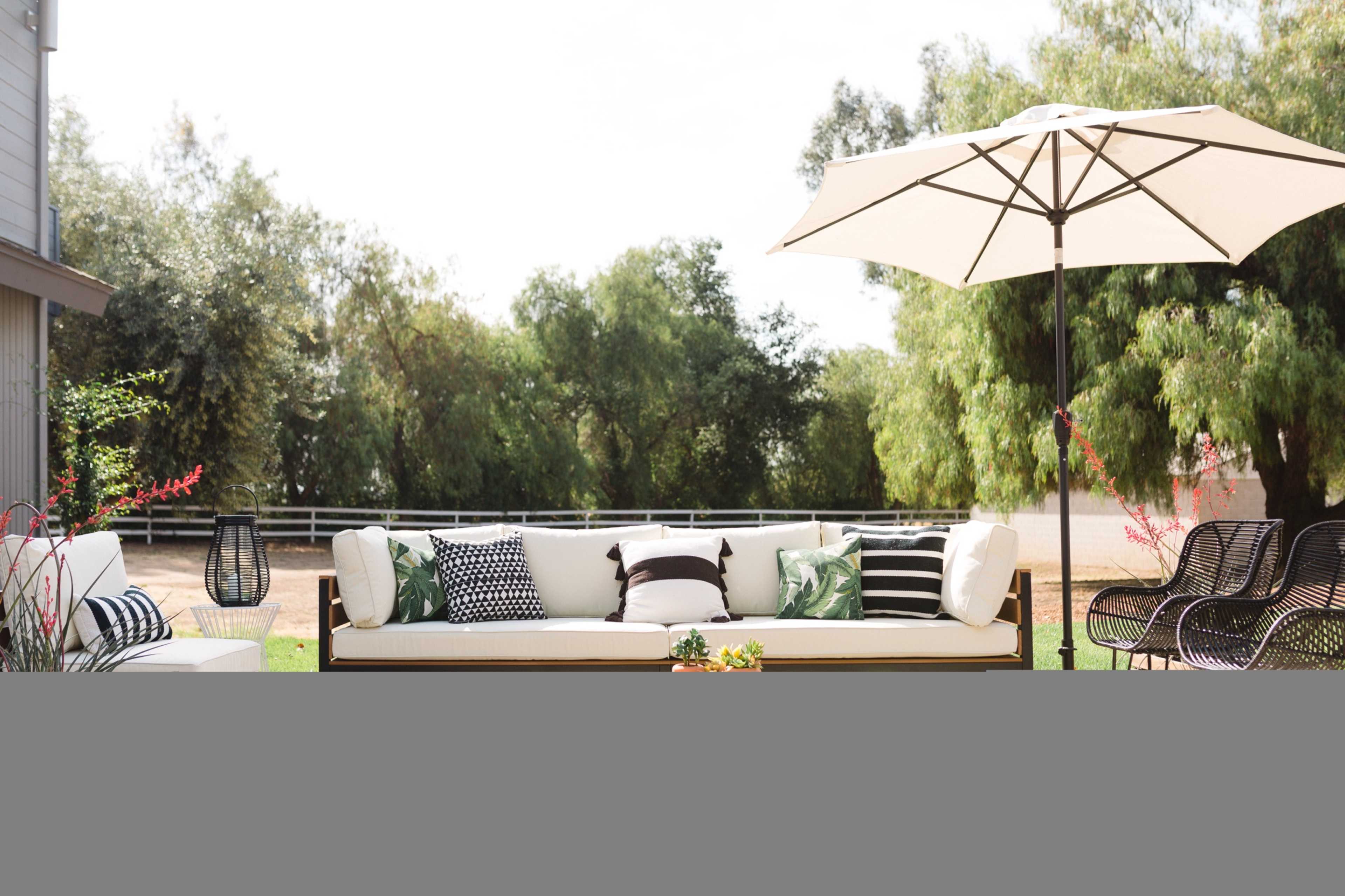 A patio area features a large sofa with various patterned cushions, surrounded by greenery and a large umbrella providing shade.