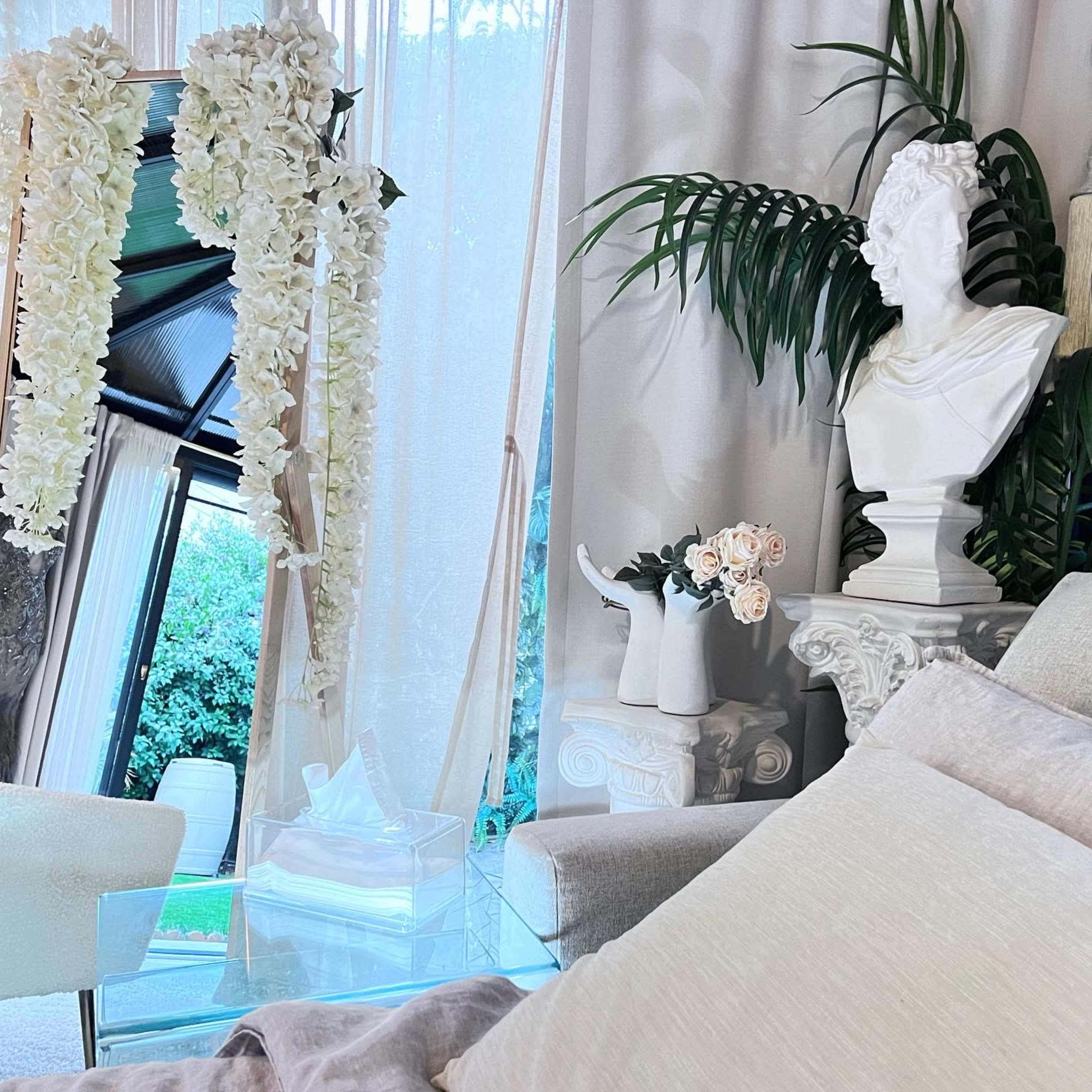 The scene features a modern living space with a bust sculpture, floral decorations, and a glass table amidst light-colored furnishings.
