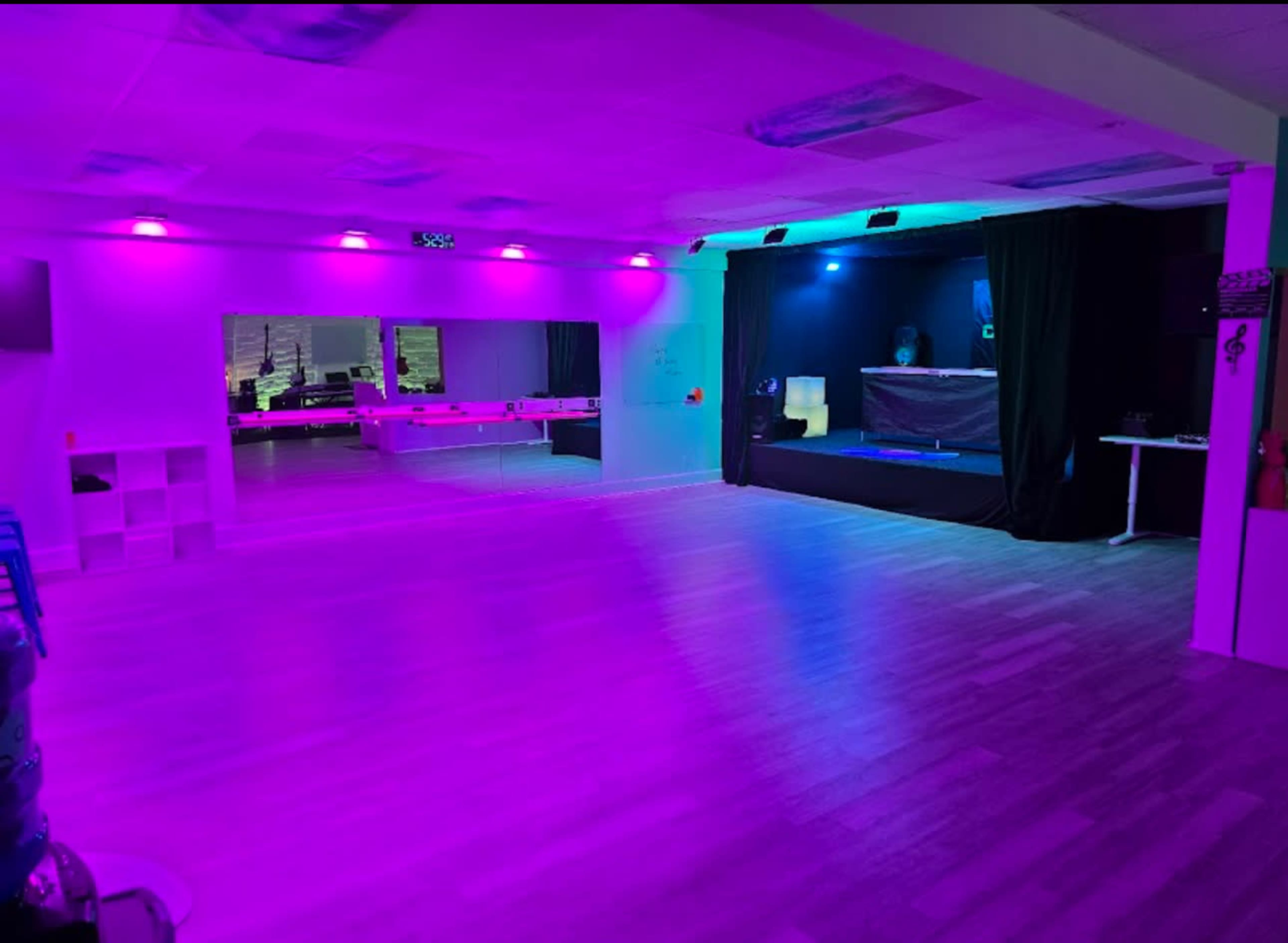 The image shows a brightly lit dance studio or event space with purple and blue lighting, featuring a stage area in the back and a large mirror on one side.