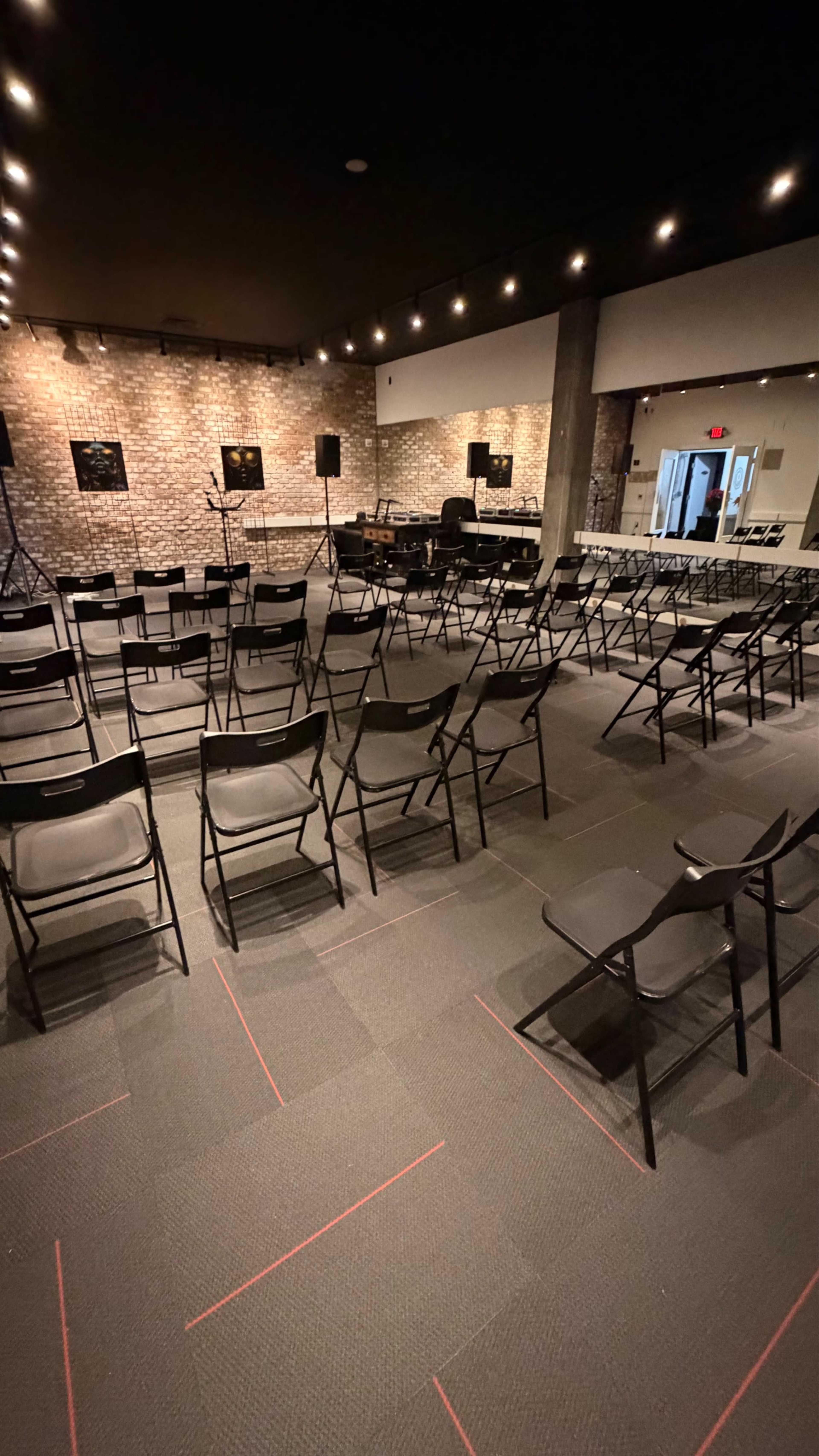 The image shows an empty event space with rows of black folding chairs facing a stage area, illuminated by dim lighting and featuring brick walls in the background.