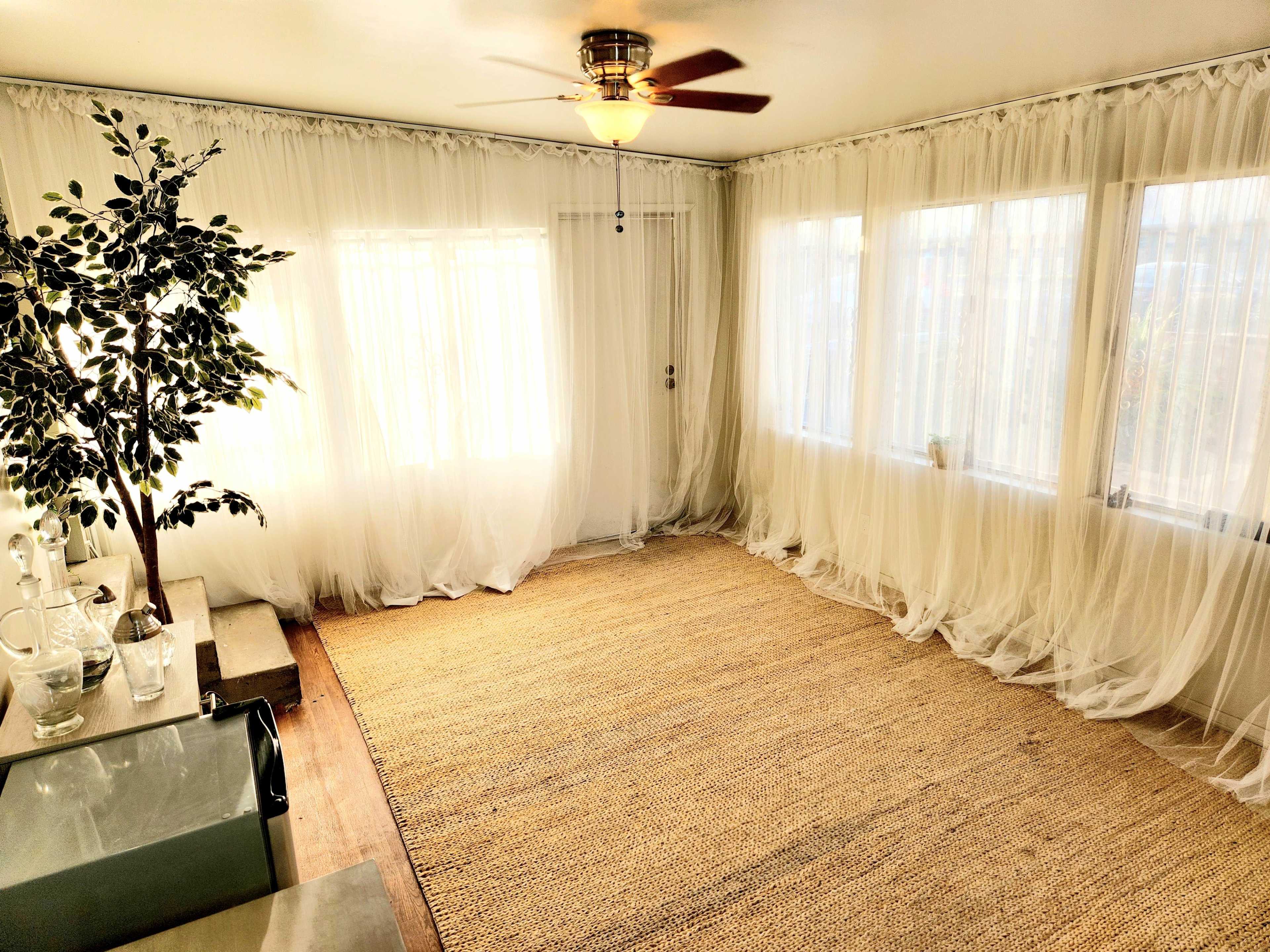 A sunlit room features sheer white curtains, a ceiling fan, a woven rug, and a potted plant in the corner.