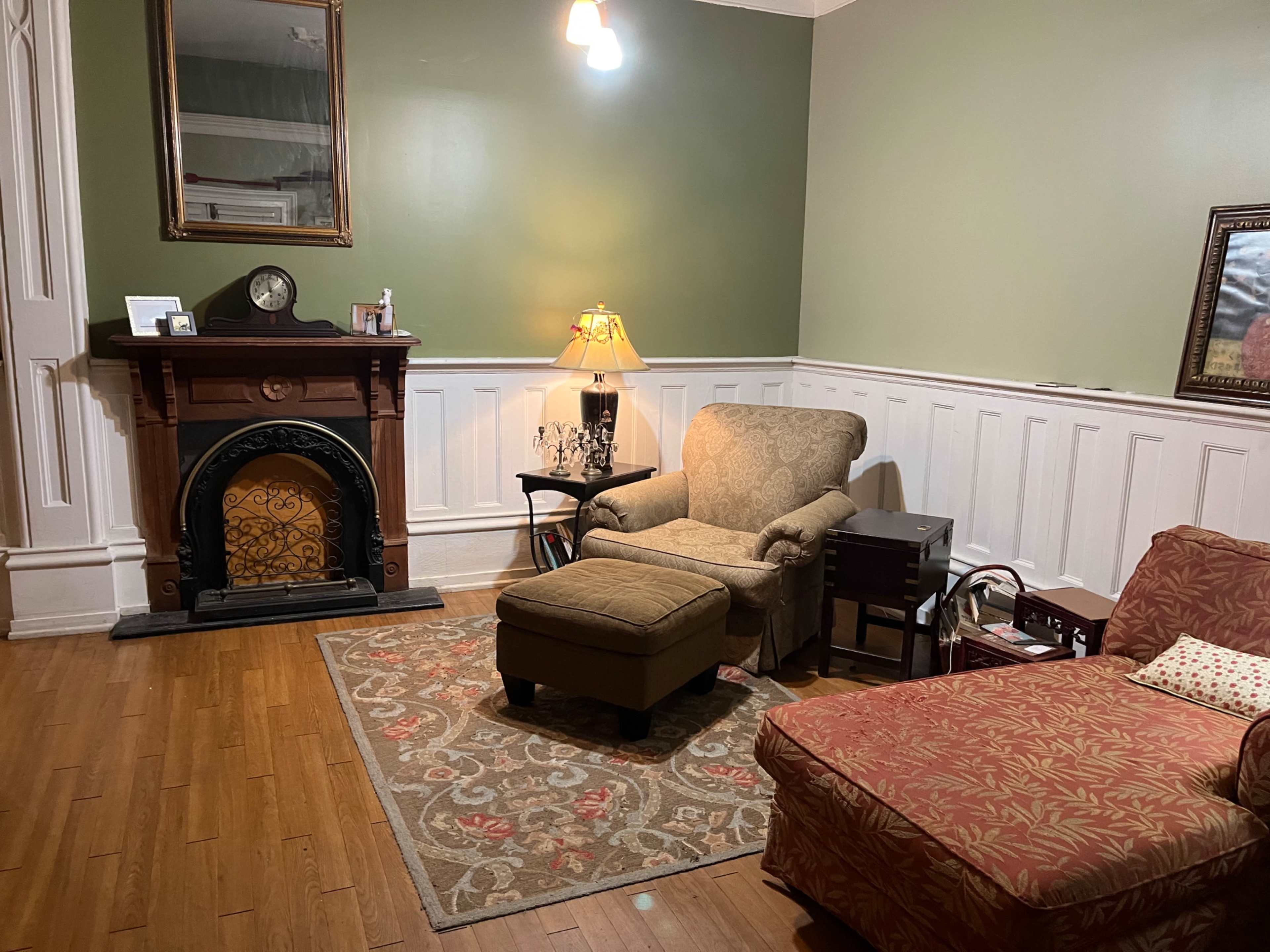 A cozy living room featuring a fireplace, a patterned area rug, an armchair, an ottoman, and a small side table with a lamp.