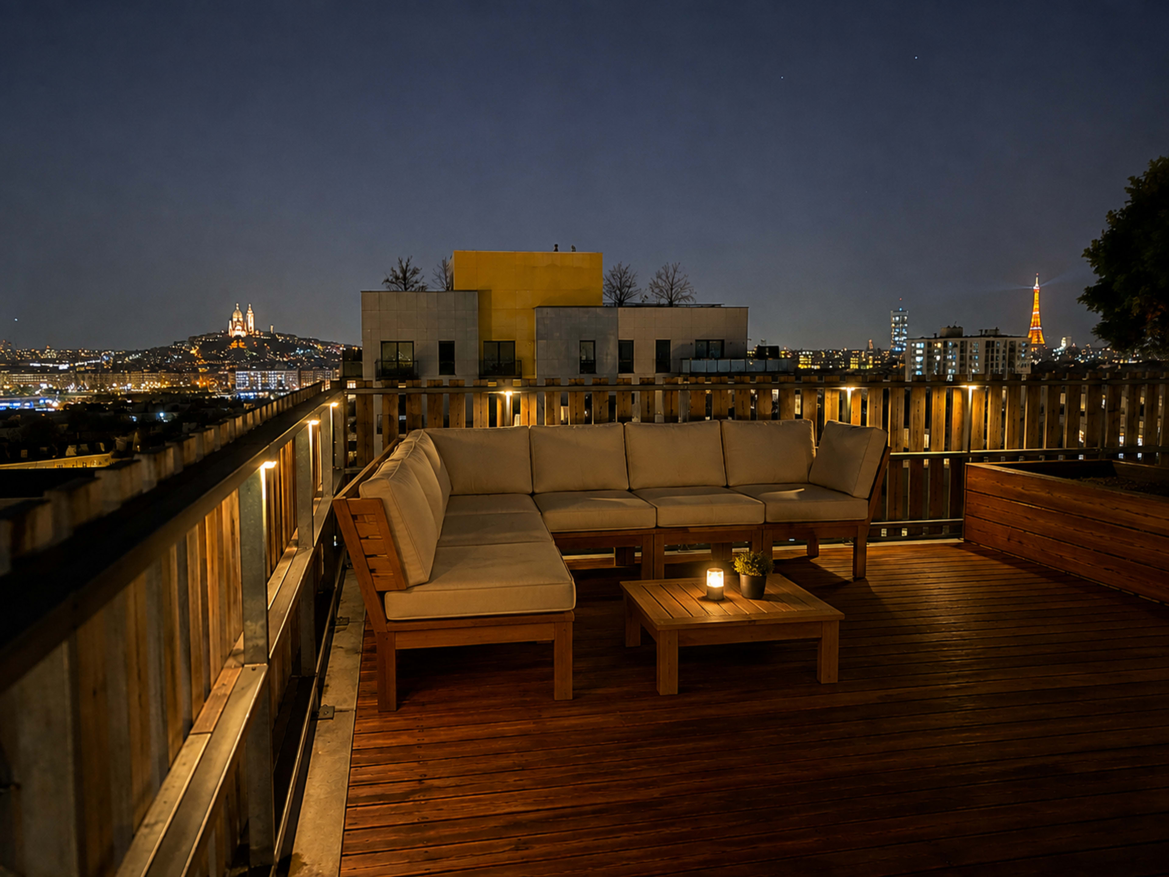 The image shows a rooftop seating area with a wooden deck, featuring a large sectional sofa and a small table, overlooking a city skyline lit at night.