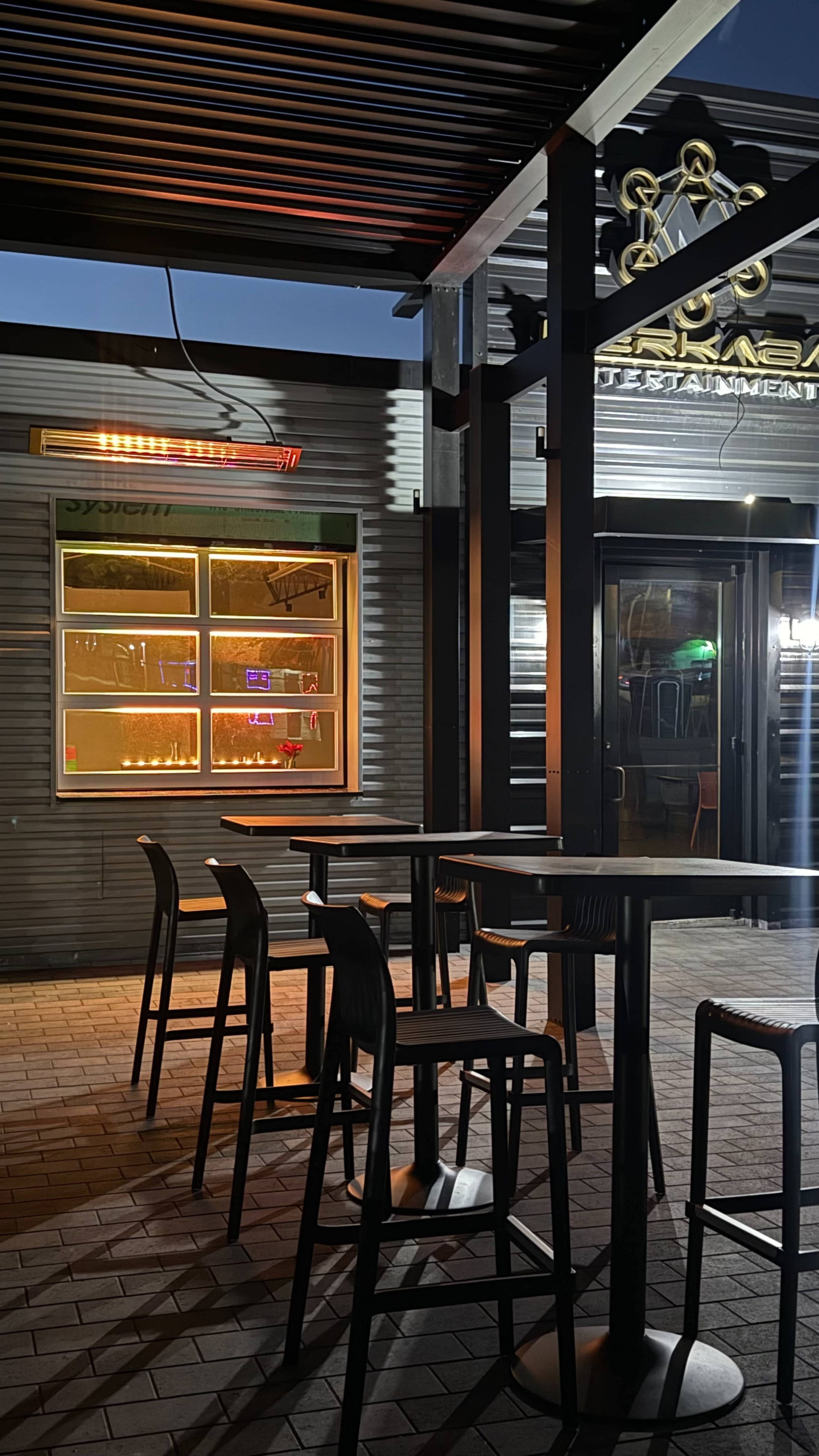 The image shows a dimly-lit outdoor seating area with several tall tables and chairs in front of a restaurant, illuminated by warm lights.