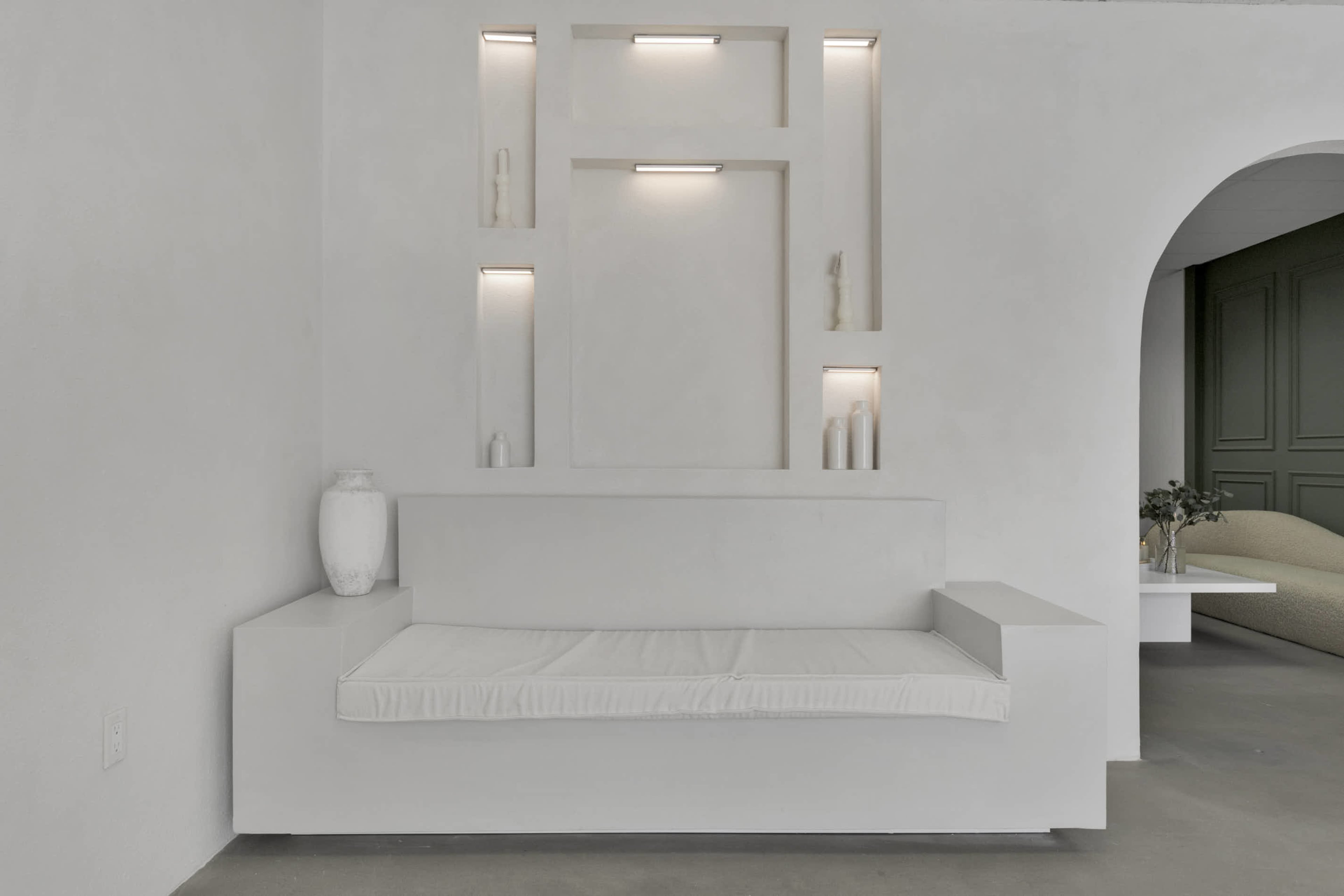 A minimalist white bench is positioned against a wall with built-in shelving, illuminated by recessed lights.