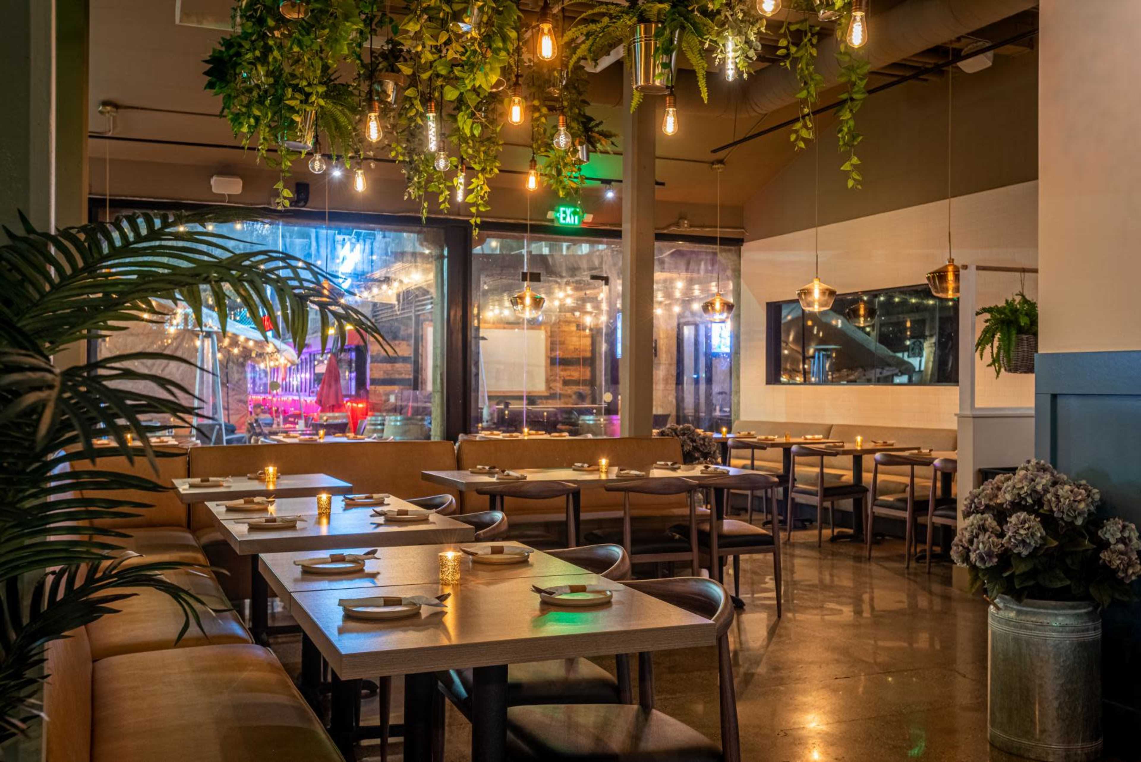 A modern restaurant interior with wooden tables set for dining, surrounded by decorative plants and ambient lighting.
