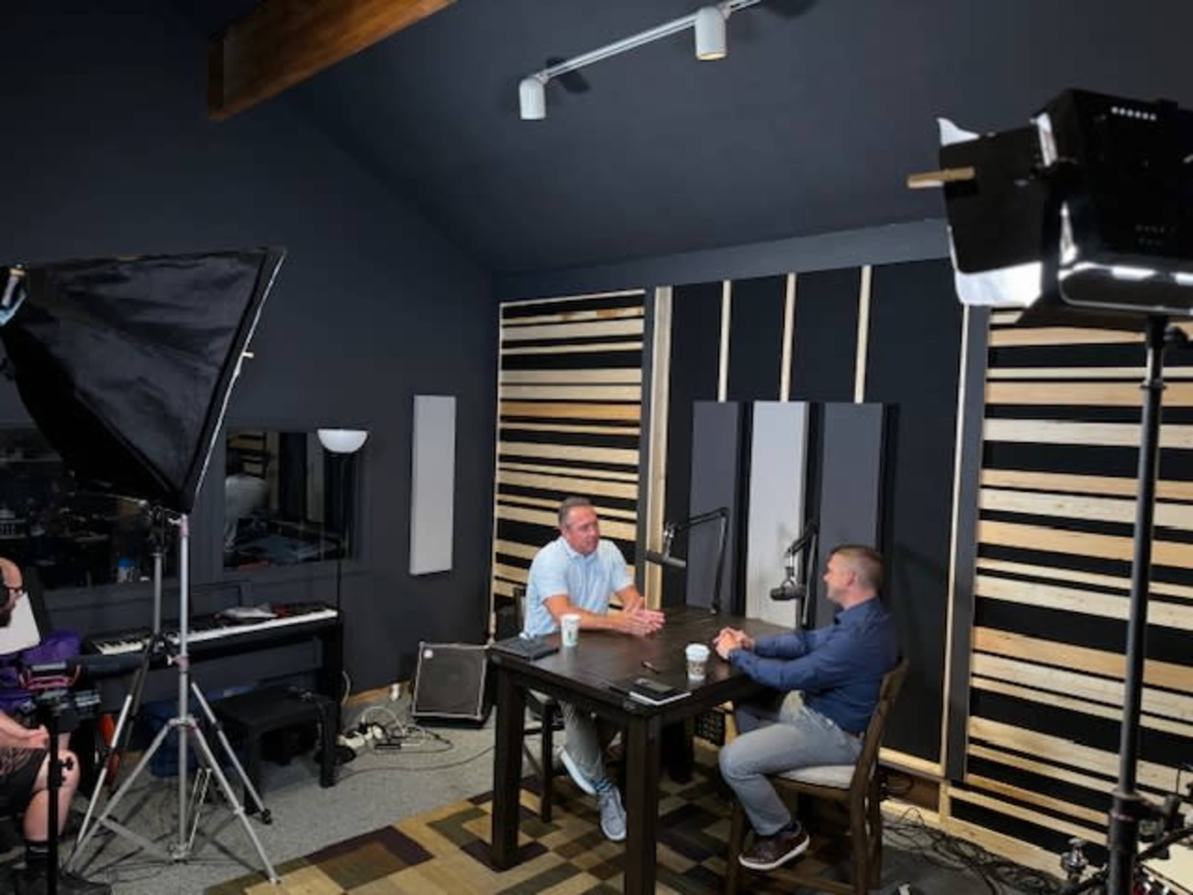 Two men are seated at a table in a studio-like setting with wooden paneling on the walls and studio lighting equipment nearby.