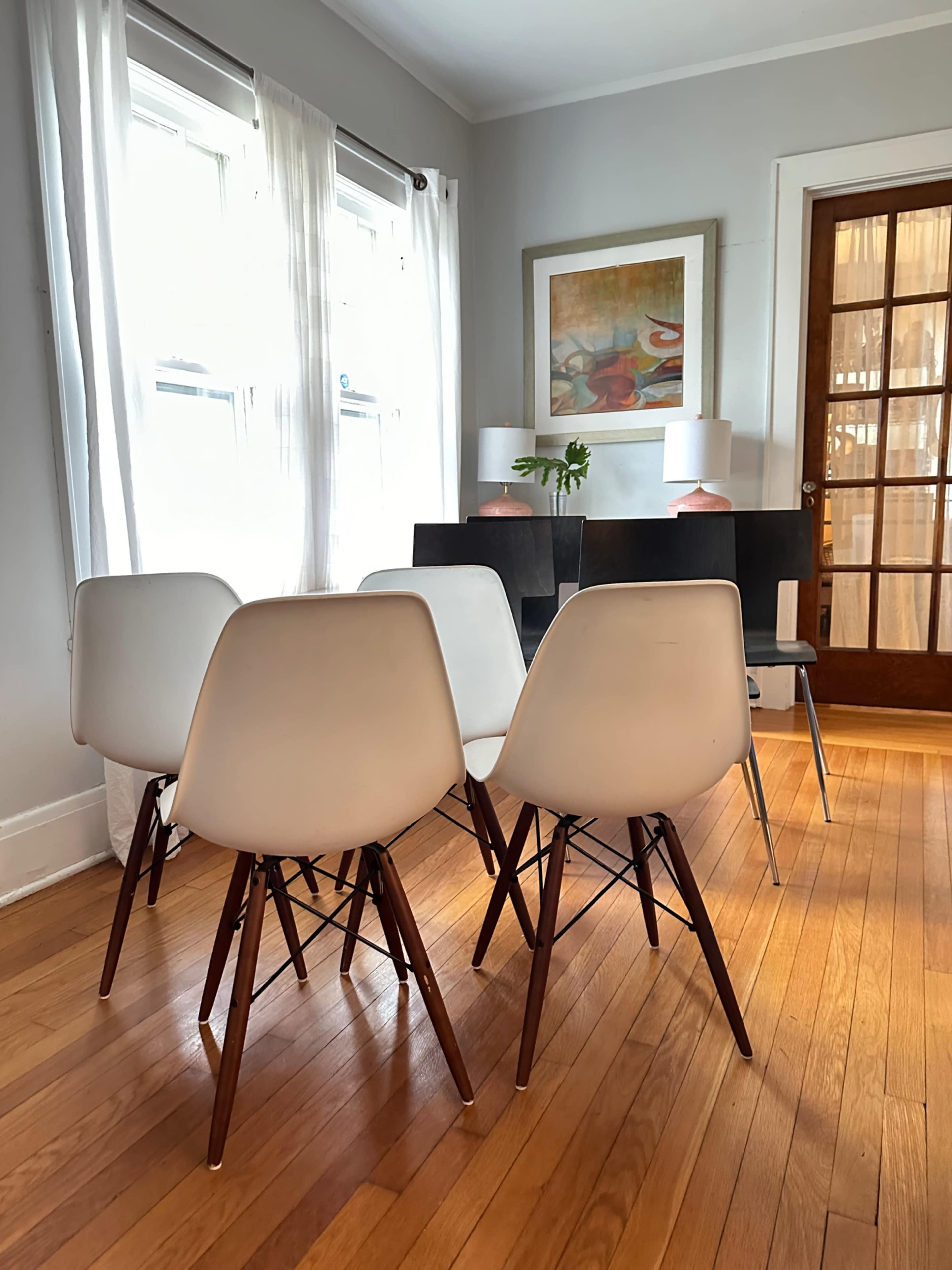 A row of white chairs with wooden legs faces a table in a sunlit room, featuring large windows and a door with glass panels.