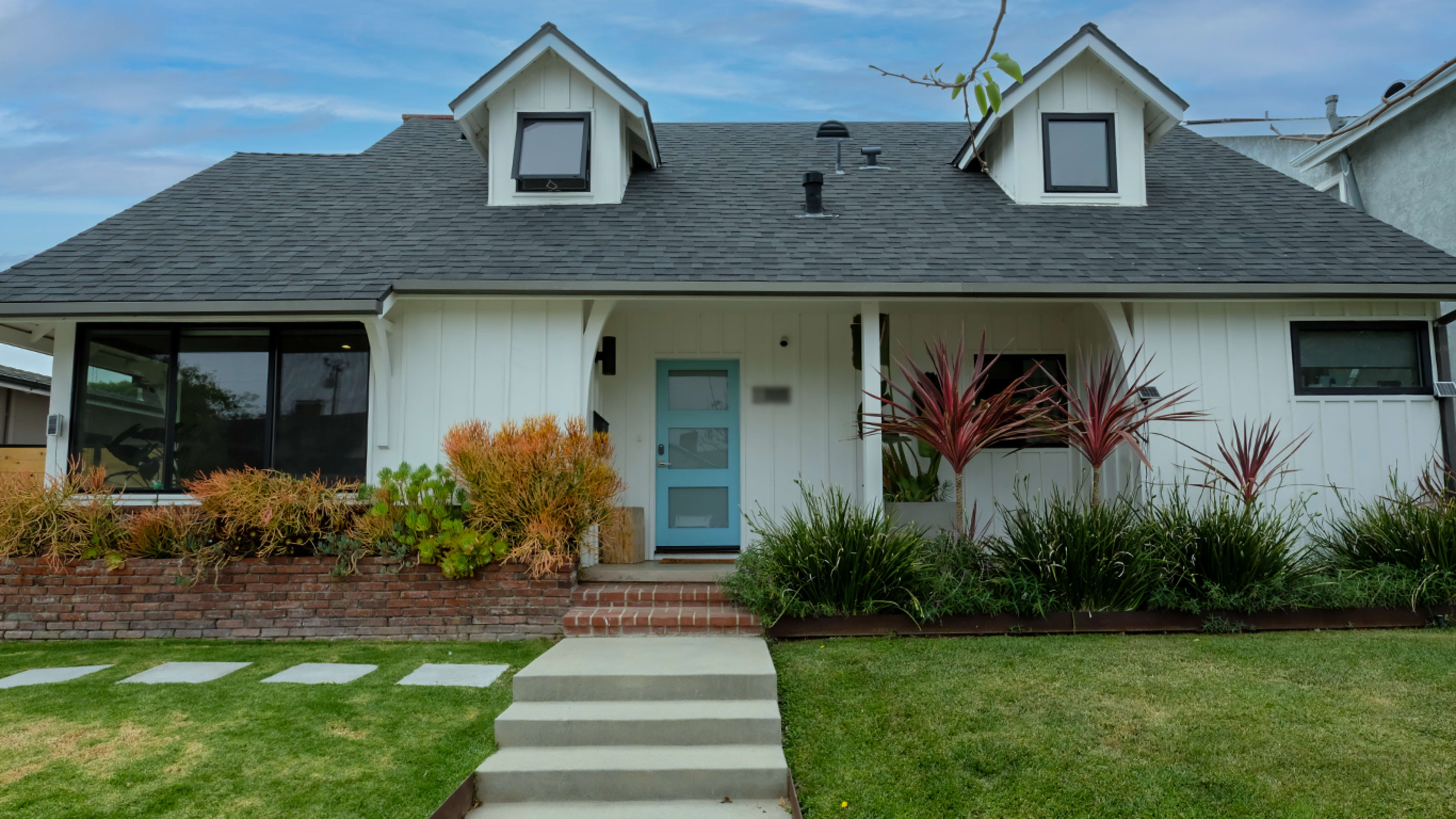 A white two-story house with a gray roof, two front dormers, and a landscaped yard featuring decorative plants.
