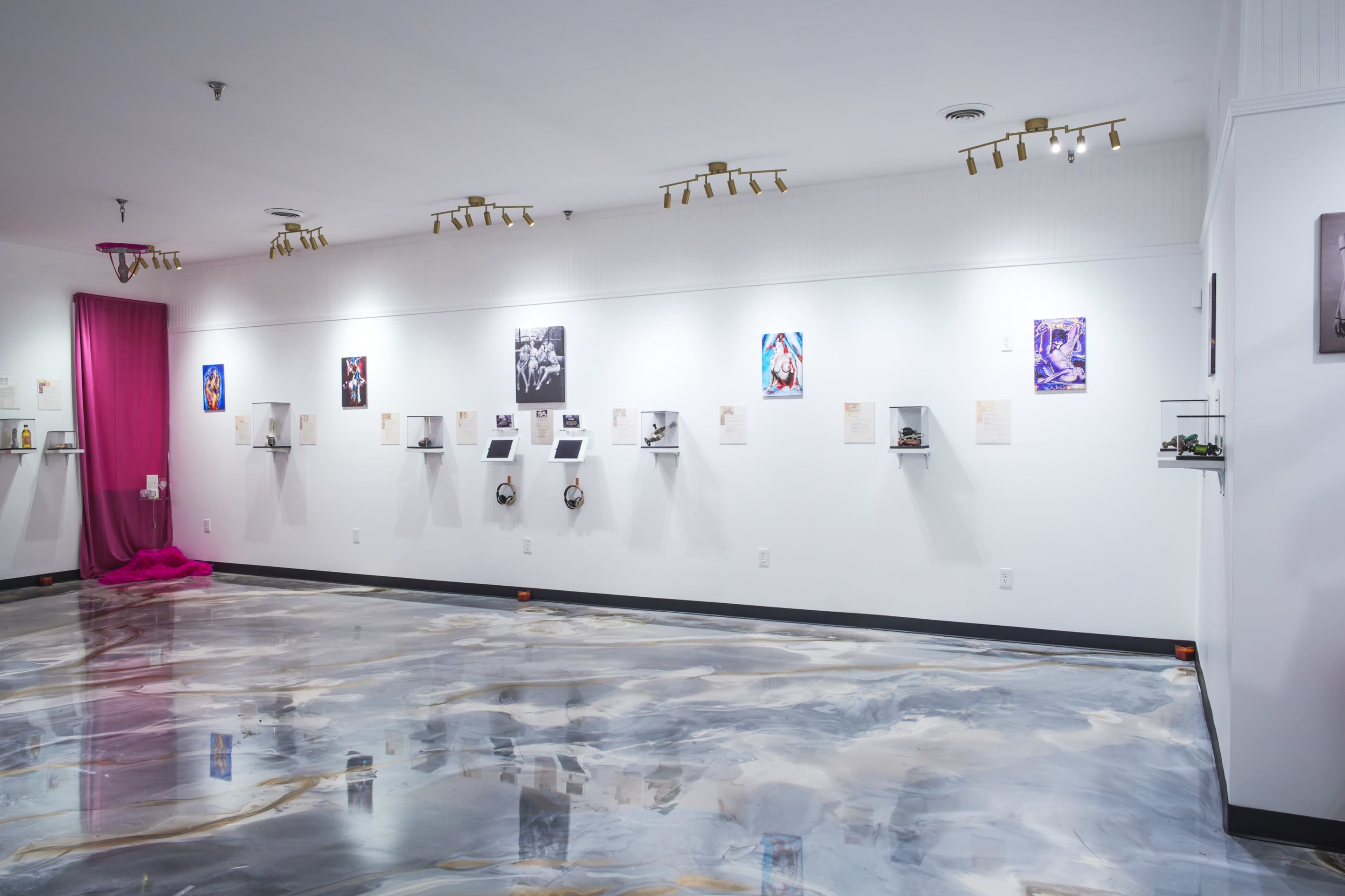 The image shows a modern art gallery with a polished floor, displaying various framed artworks on white walls and small shelves.