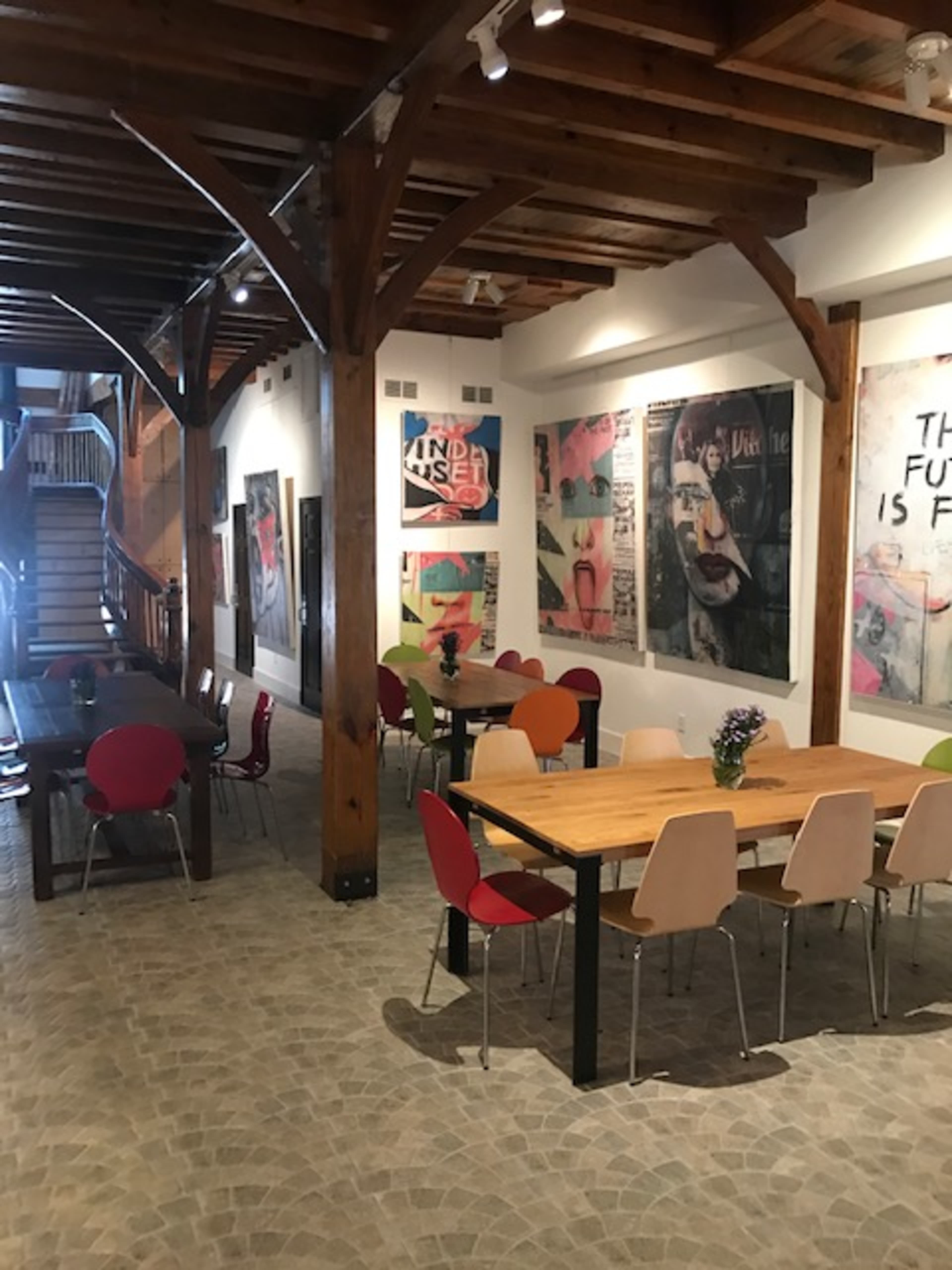 A well-lit interior of a café or community space with wooden beams, colorful chairs around tables, and large artwork on the walls.