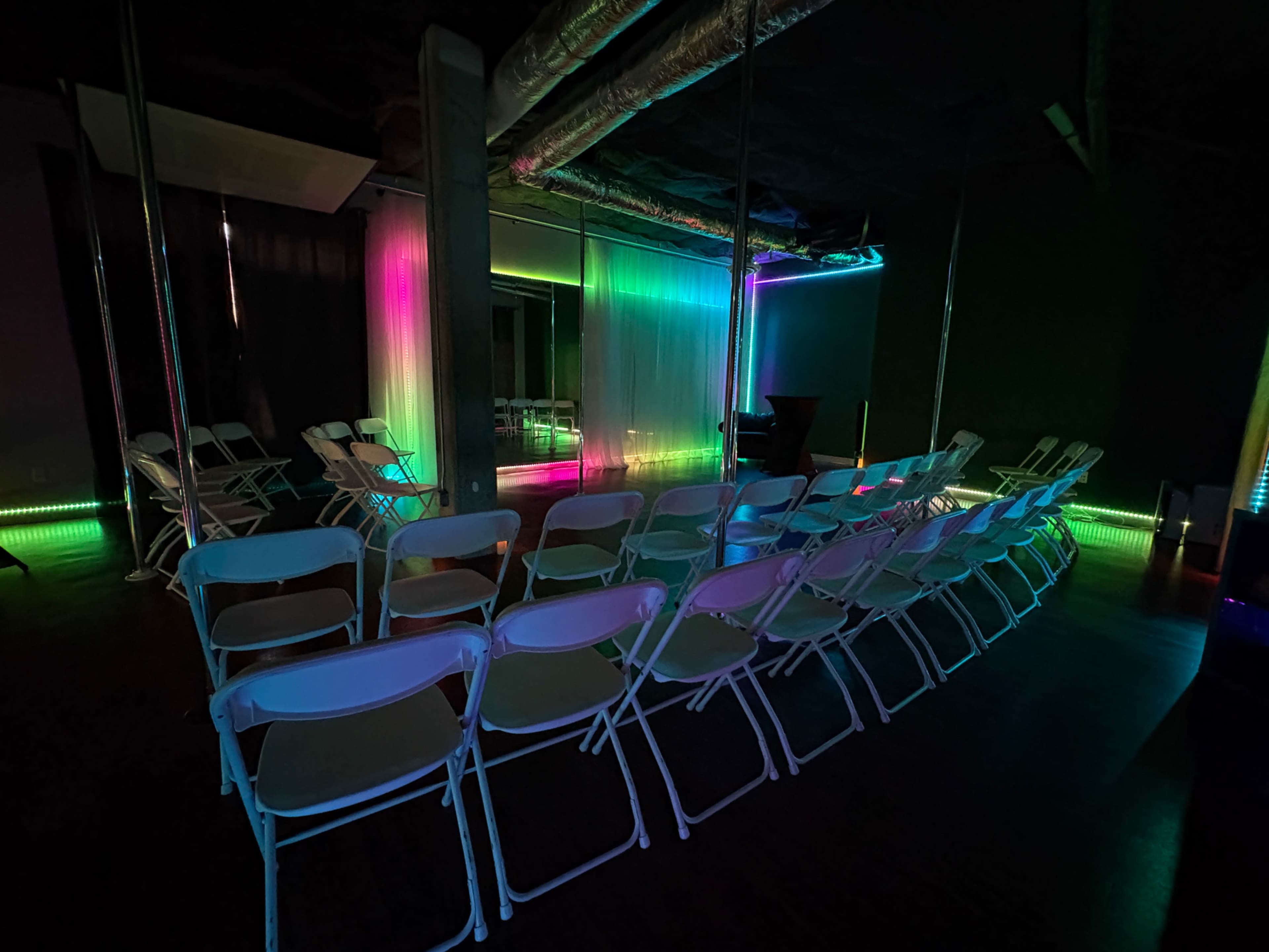 A row of white folding chairs faces a brightly lit stage with colored LED lights illuminating the walls in a dimly lit room.
