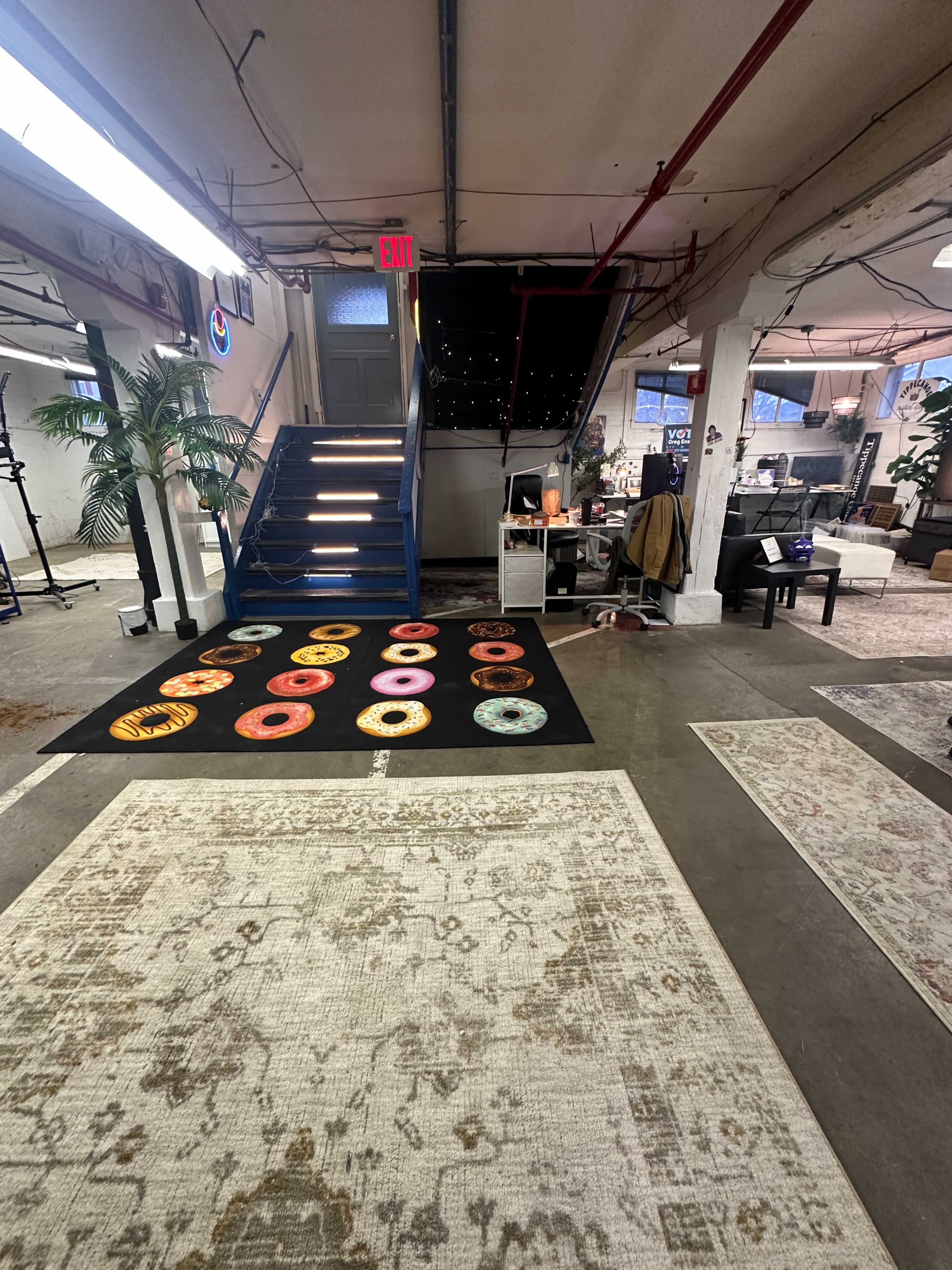 The image shows a spacious room with various rugs on the floor, a staircase leading to an upper level, and an assortment of furniture and plants.