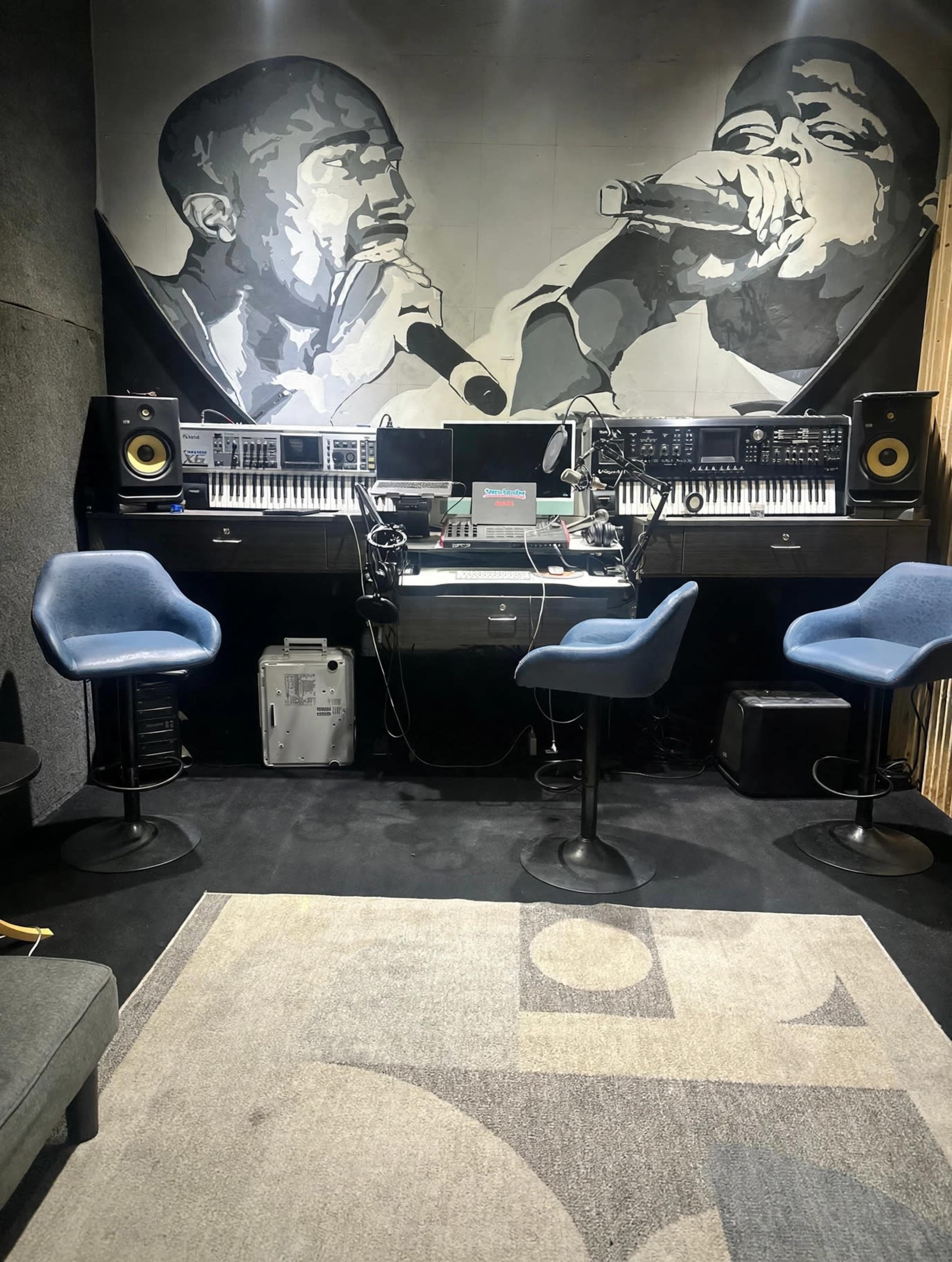 The image shows a music studio featuring two blue chairs in front of a mixing console, keyboards, and a mural of two artists on the wall.