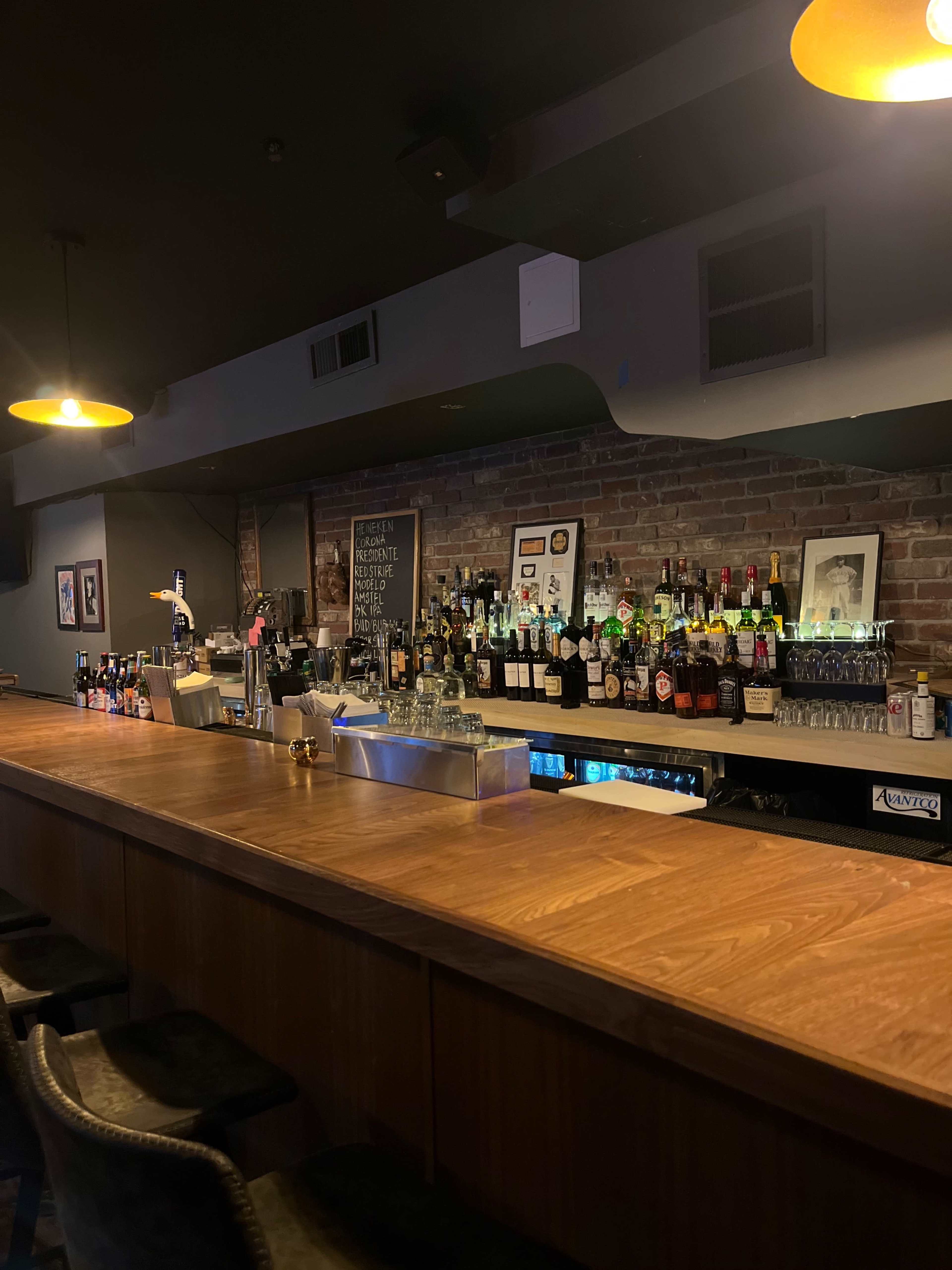 A bar with a wooden counter and a variety of liquor bottles arranged on shelves against a brick wall.