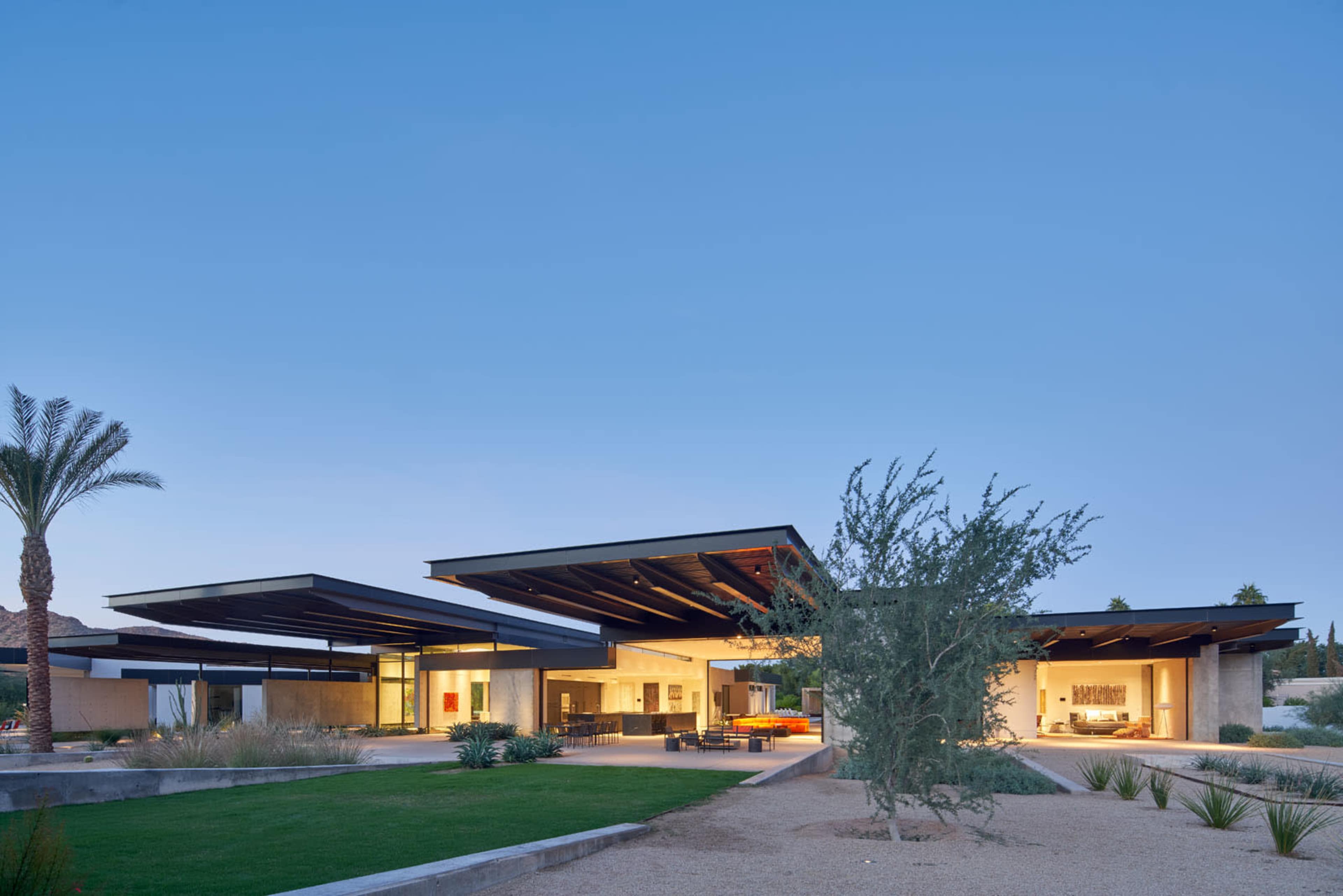 A modern house with flat roofs and large glass windows is set against a clear blue sky, surrounded by landscaped greenery and desert plants.