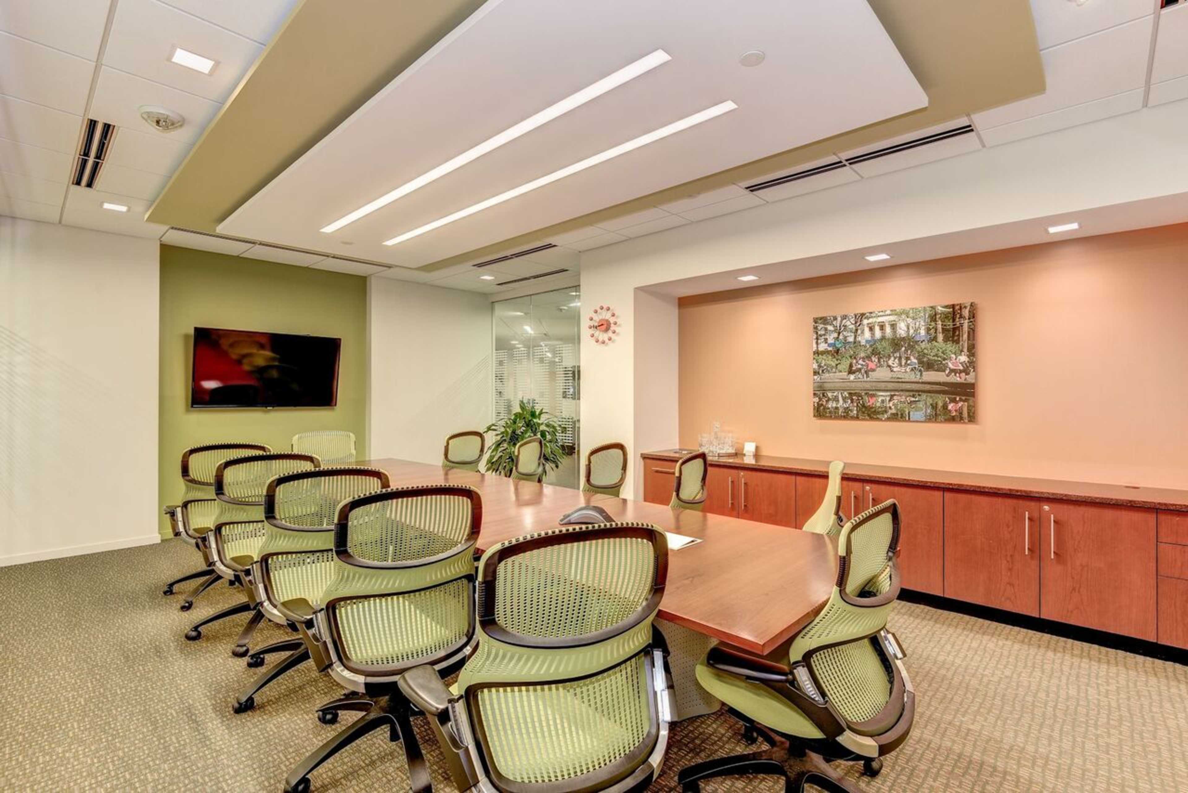 A conference room with a large wooden table surrounded by ergonomic green chairs, a television on the wall, and a decorative picture.