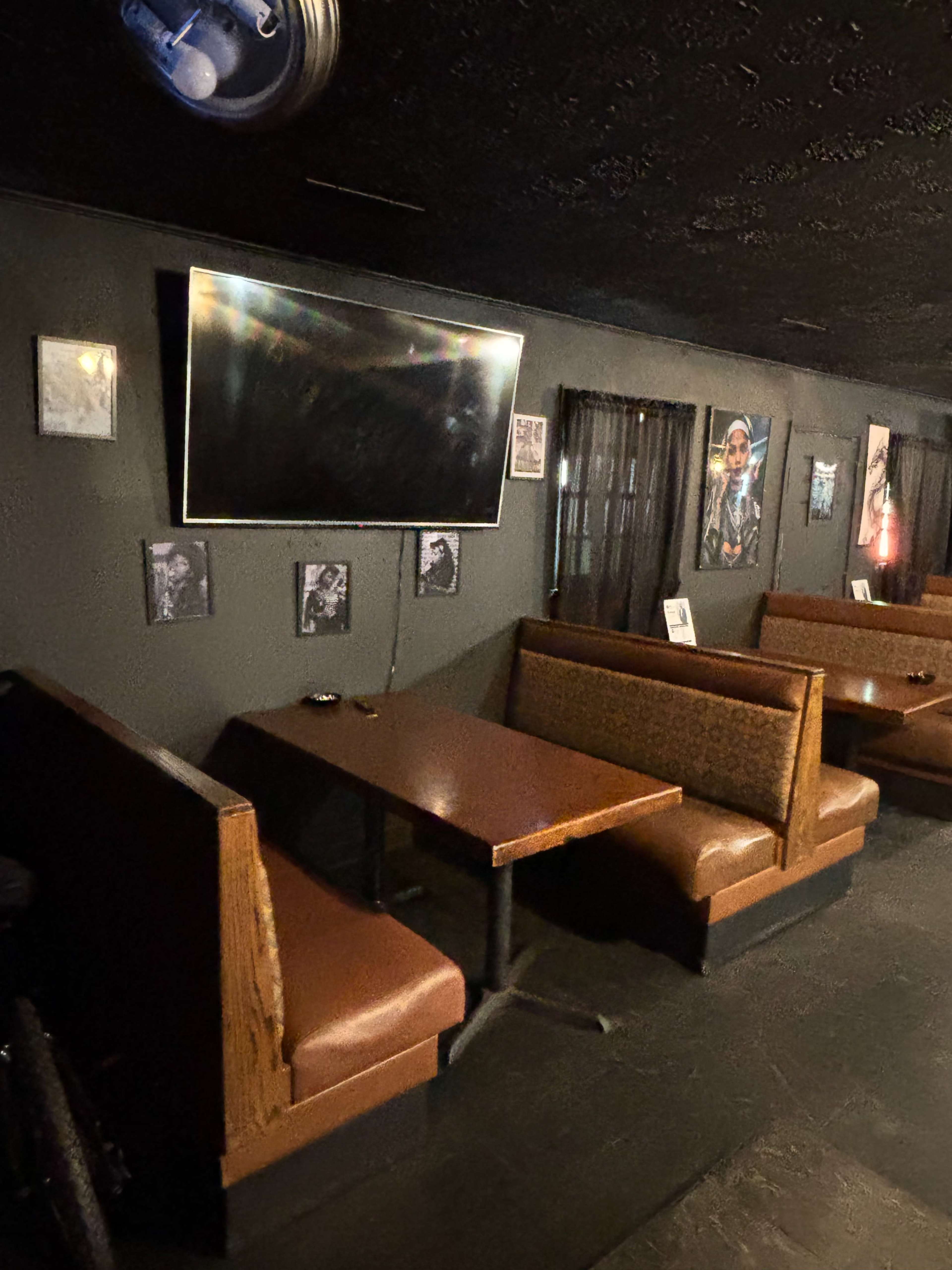 The image shows a dimly lit seating area in a restaurant or bar, featuring a wooden table with two padded benches and a wall-mounted television above it, surrounded by framed photographs.