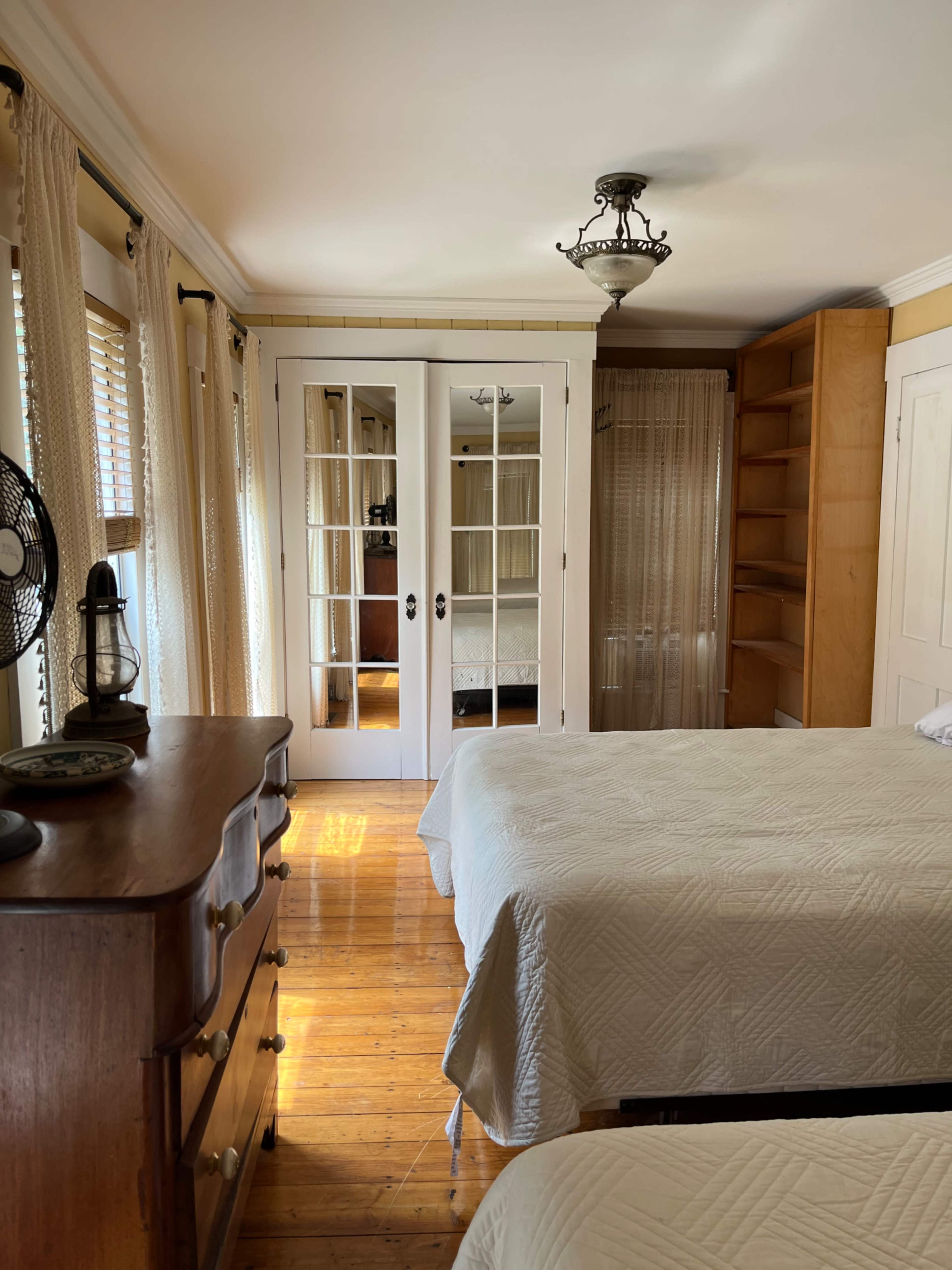 A bright bedroom with two beds, a wooden dresser, and a set of double doors leading to another room.