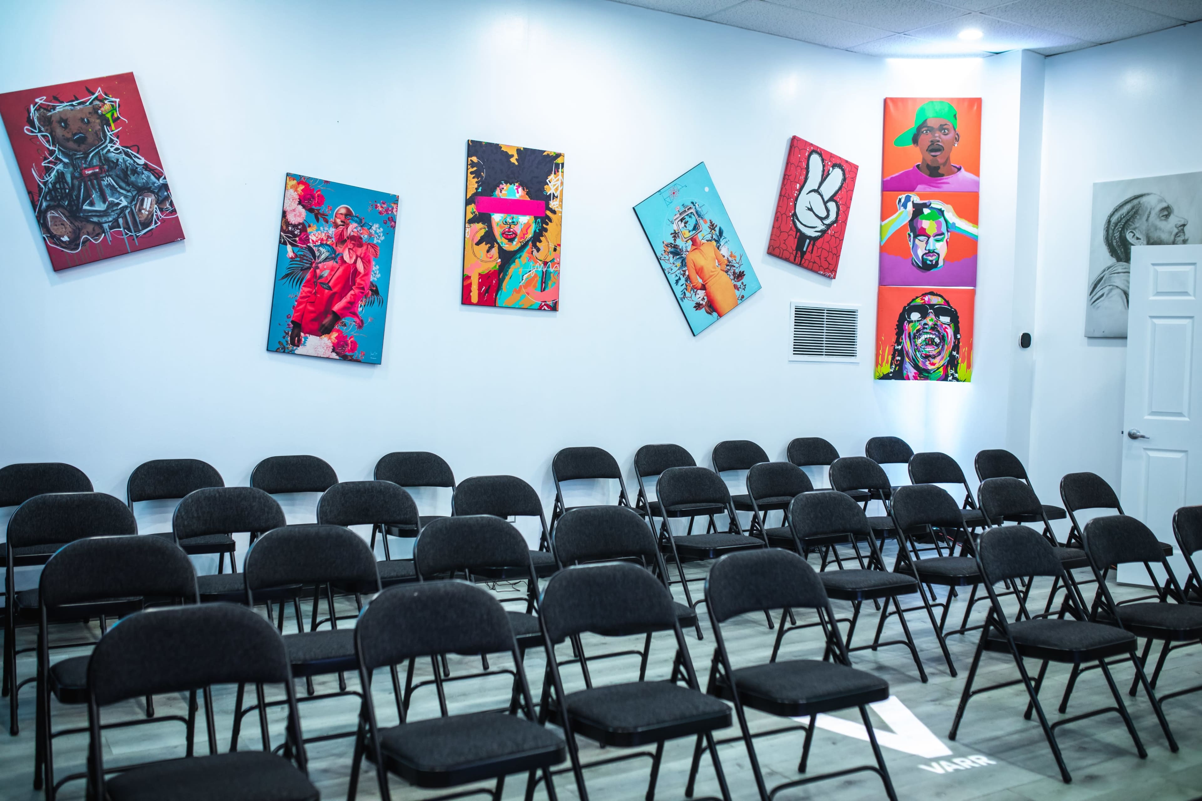 A room with several black folding chairs arranged in rows and vibrant, colorful artwork displayed on the walls.