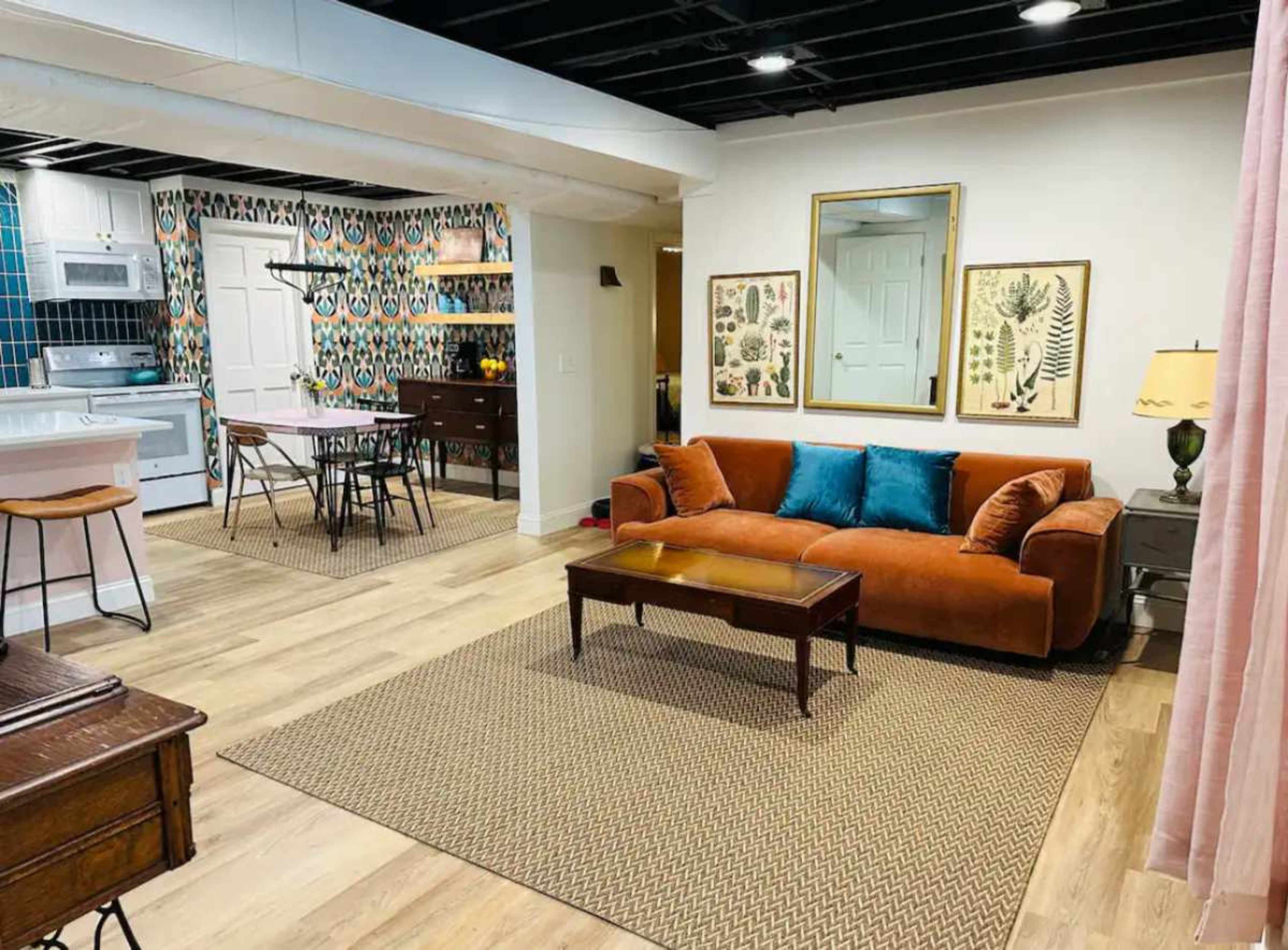 The image shows a cozy, modern living area with an orange couch, a coffee table, and wallpapered walls featuring a patterned design, connected to a kitchen and dining area.
