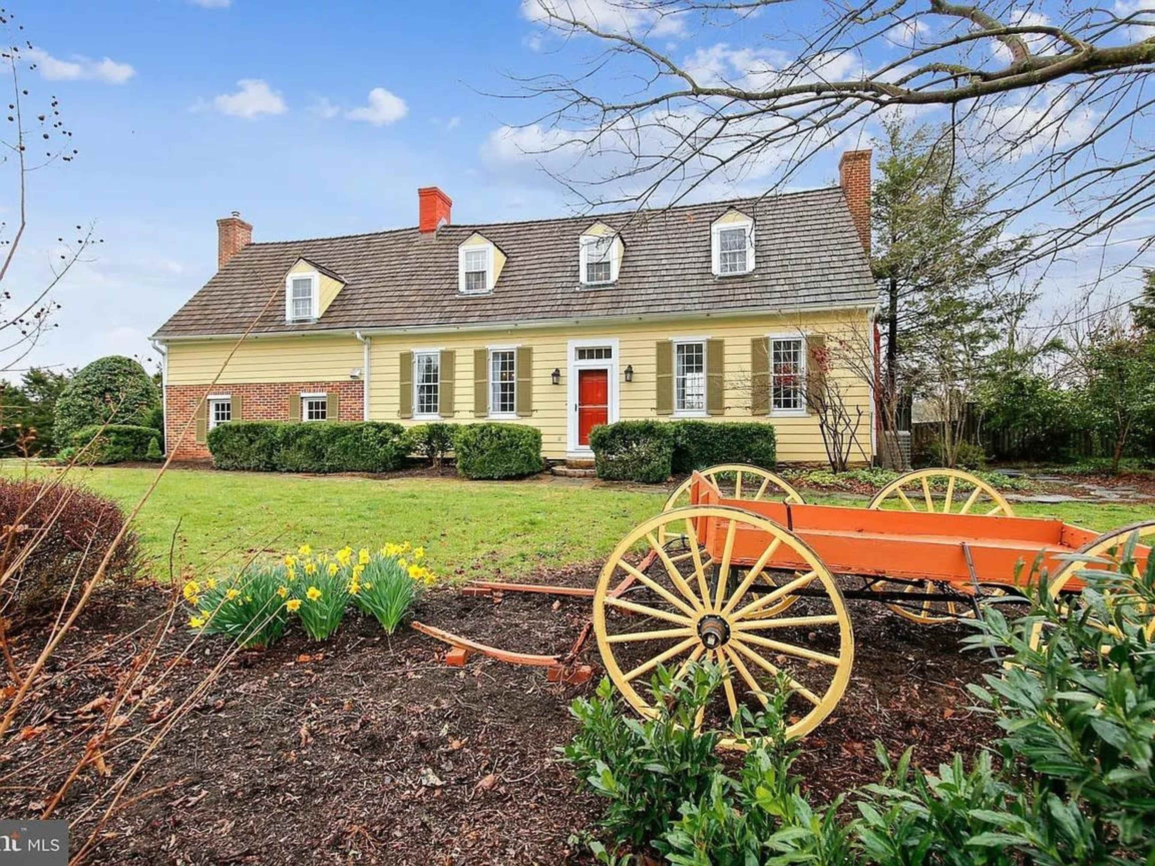 The image shows a yellow two-story house with a gabled roof and a red front door, surrounded by a grassy lawn and a decorative orange wagon placed in front of it.
