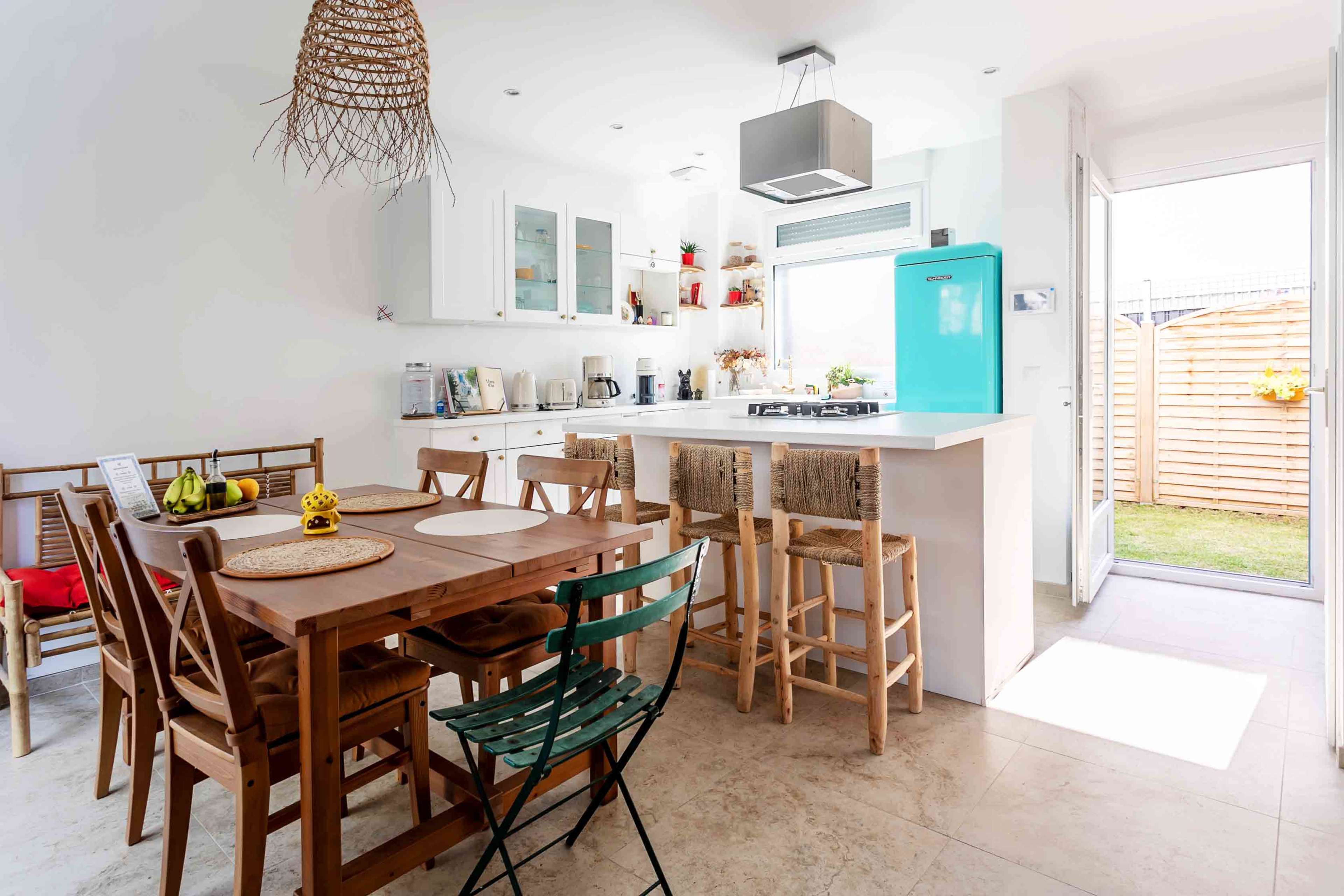 A modern kitchen features a wooden dining table with six chairs, a turquoise refrigerator, and an island with bar stools, alongside a view of a garden through the glass door.