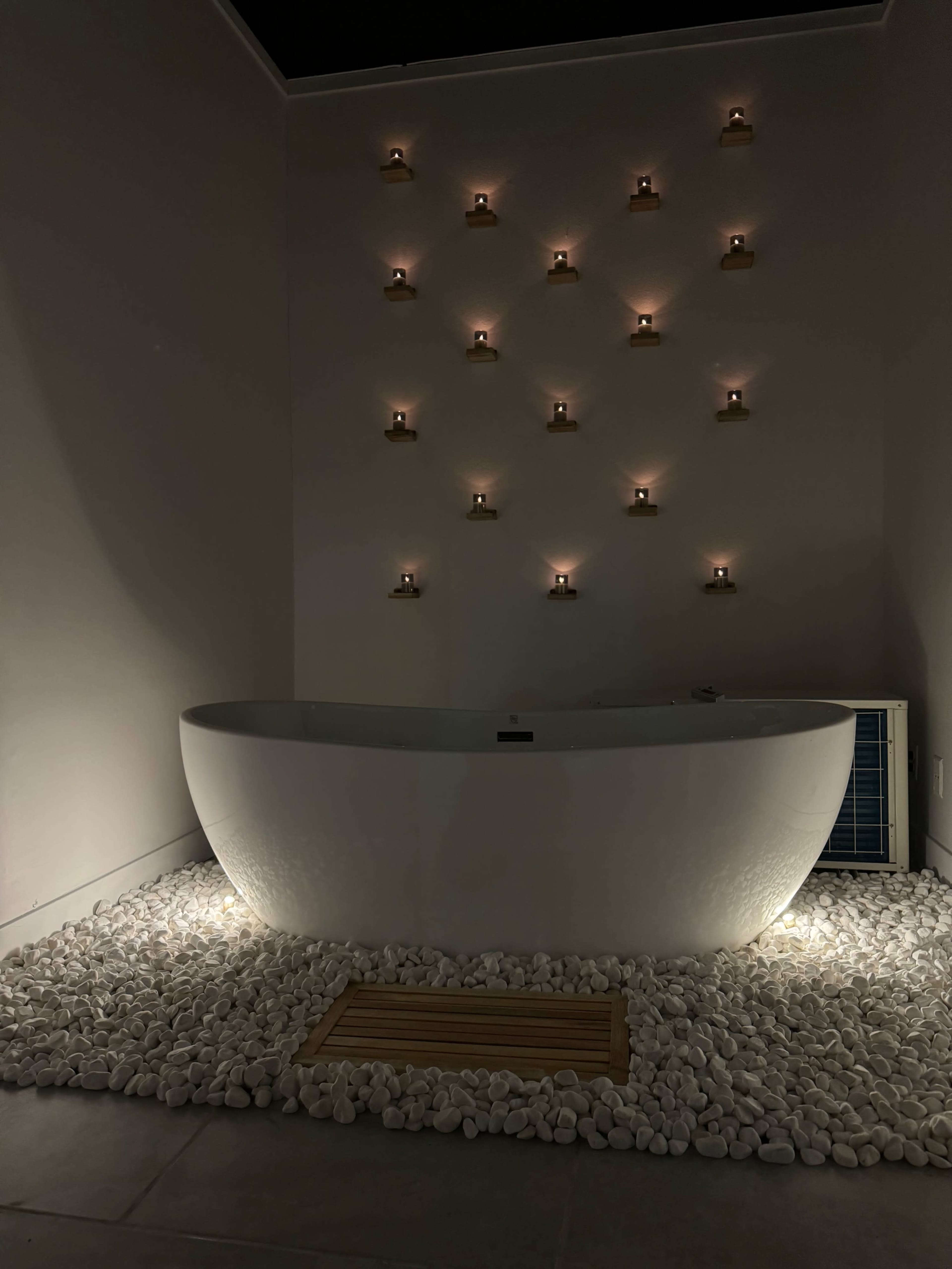 A modern bathroom features a freestanding bathtub surrounded by white pebbles, with candles arranged on the wall above.