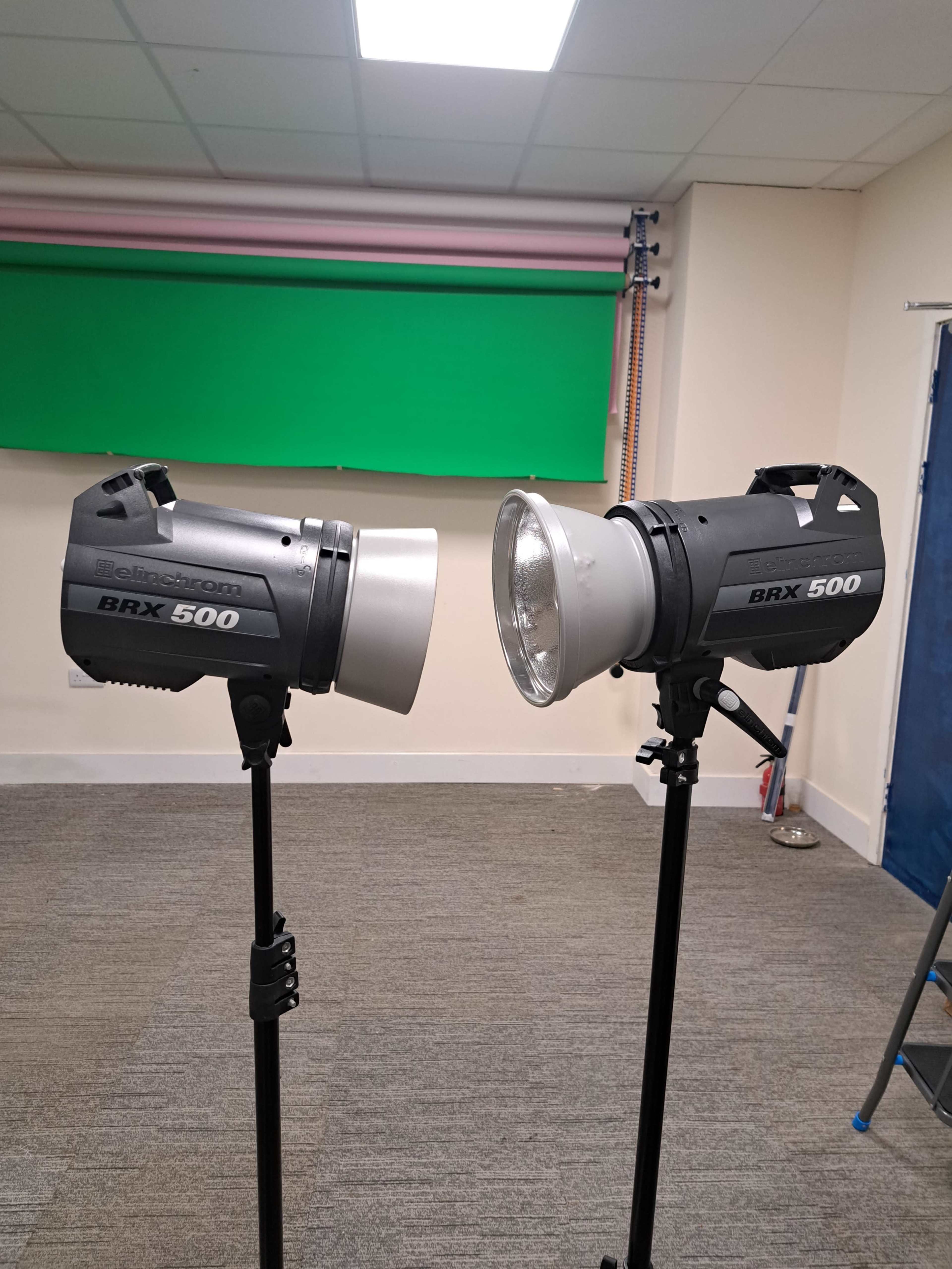 Two studio lighting equipment units labeled "BRX 500" mounted on stands in a room with a green backdrop.